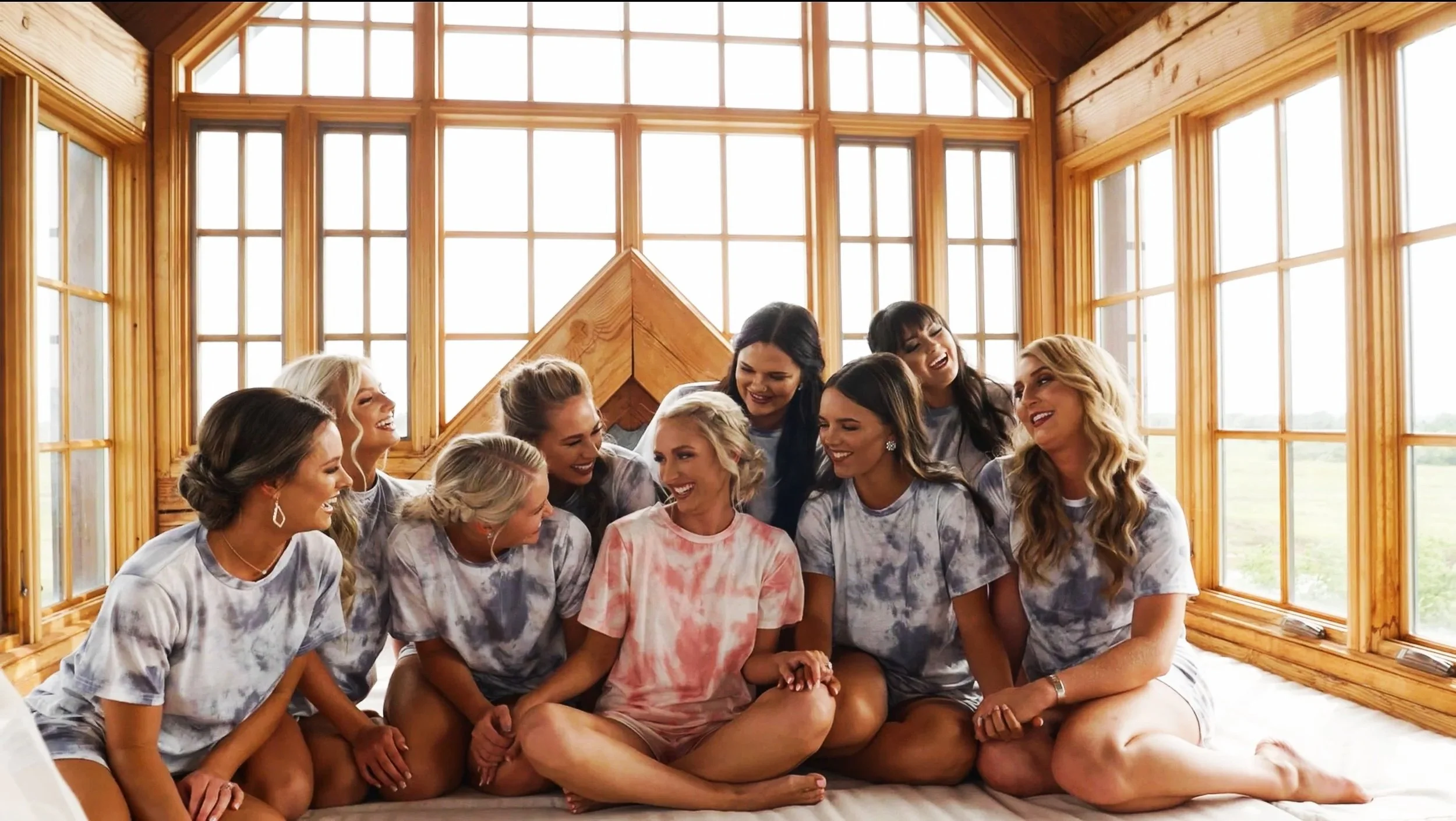 Group of women sitting on a bed in a sunlit wooden-paneled room, smiling and laughing together.