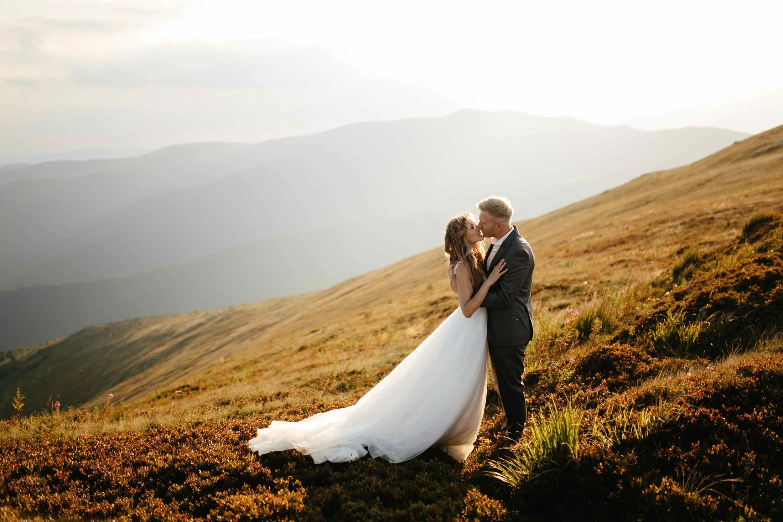 A bride and groom sharing a kiss in an open field on a hillside with mountains in the background during sunset.