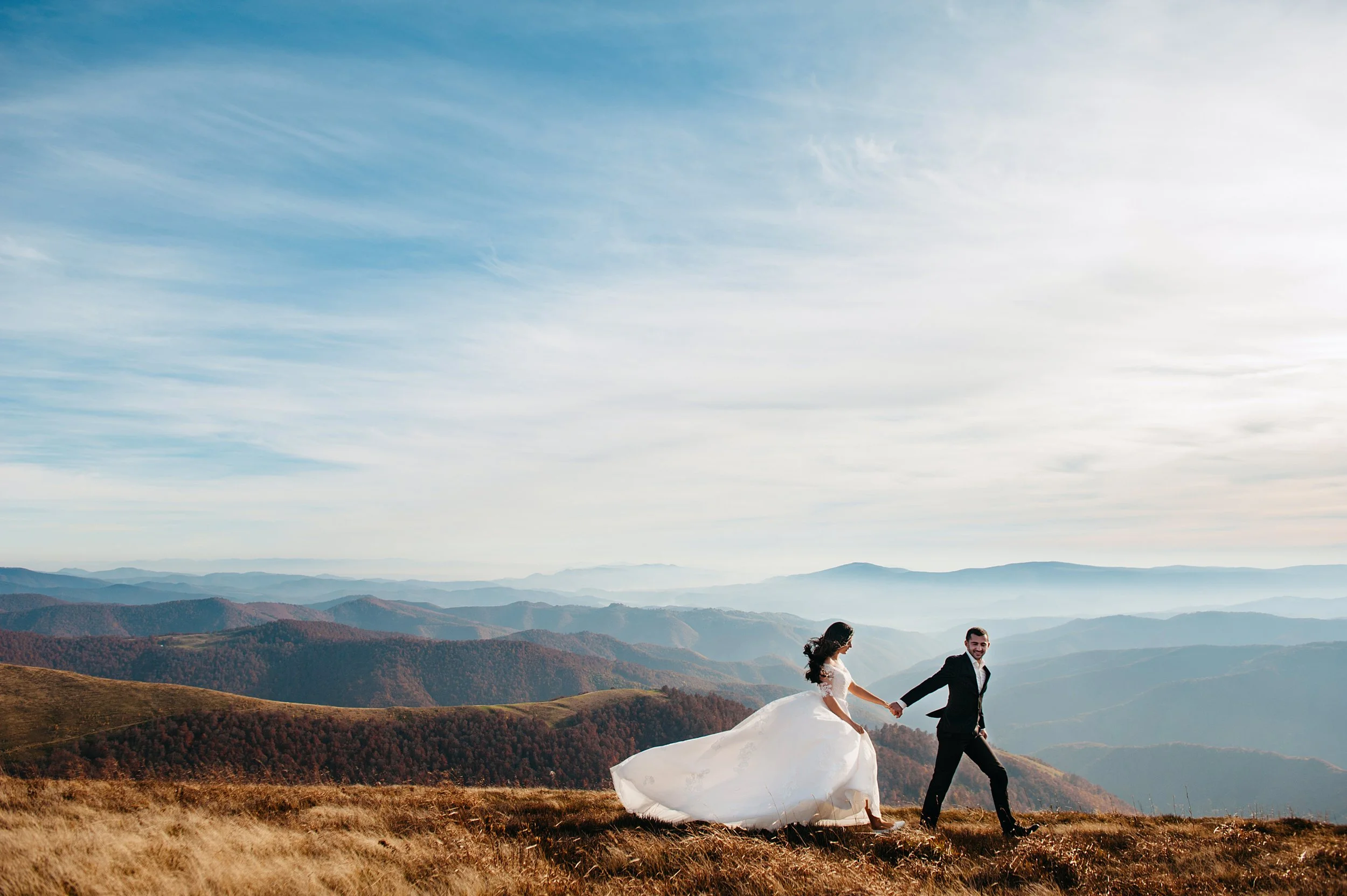 A bride and groom holding hands and walking in a mountain landscape with rolling hills and a partly cloudy sky.