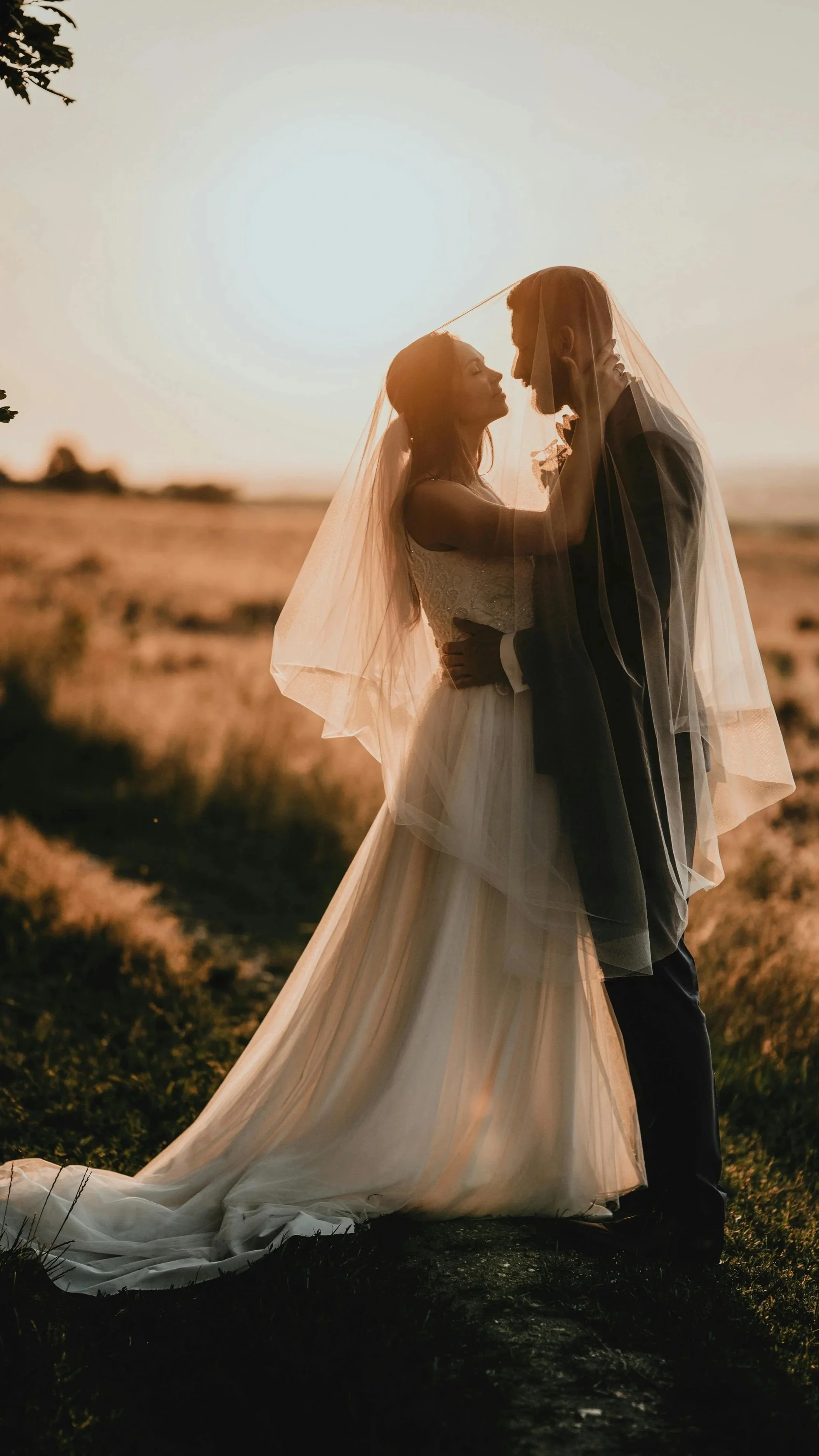 A bride and groom stand close together outdoors during sunset. The bride is wearing a wedding dress and veil, and the groom is in a suit. They are facing each other with their foreheads touching.