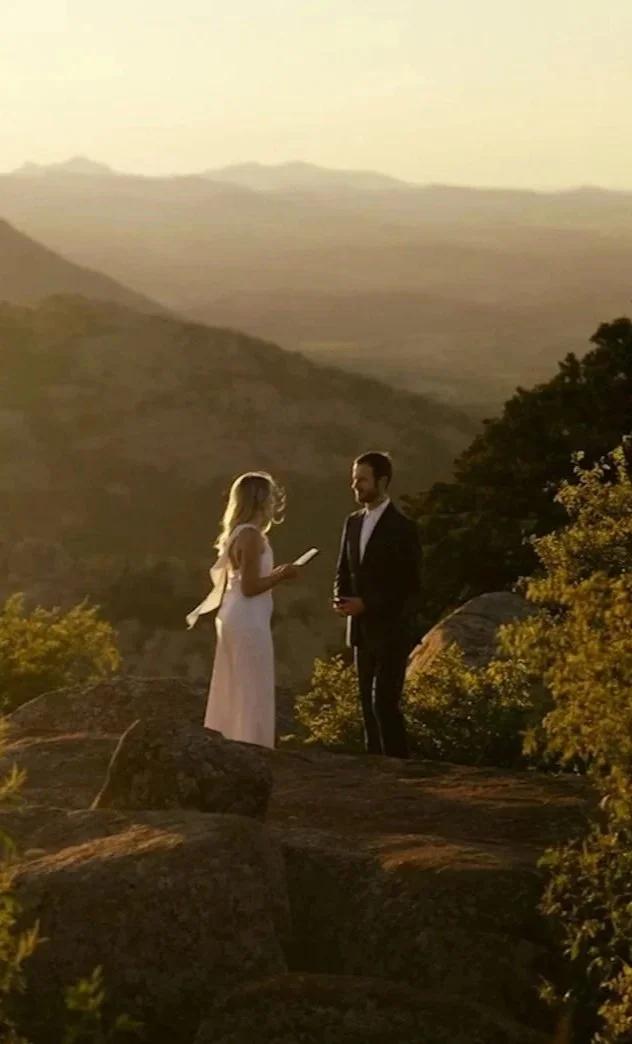 A woman in a white dress appears to be reading vows or a speech to a man in a dark suit during a sunset ceremony on a rocky hill with a mountainous landscape in the background.