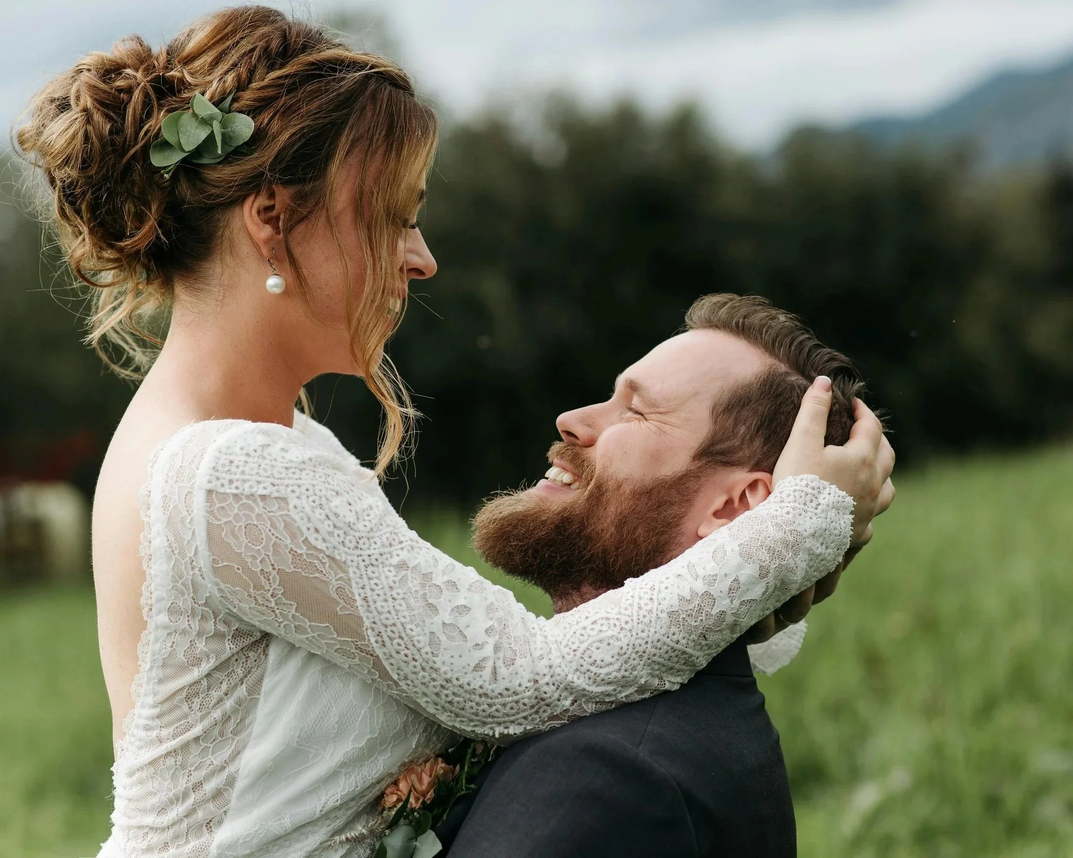 A woman in a white lace dress and a man with a beard in a dark suit embrace outdoors. She has her hands on his head, and they are looking at each other, smiling, with a blurred natural background.