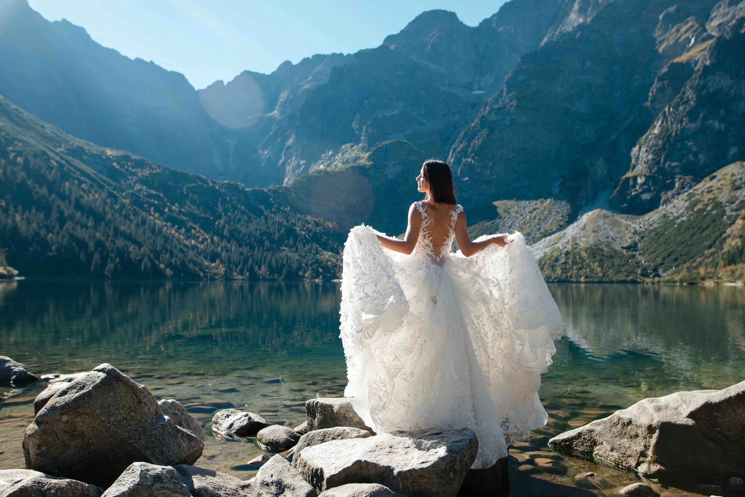 A woman in a wedding dress standing on rocks by a lake surrounded by mountains.