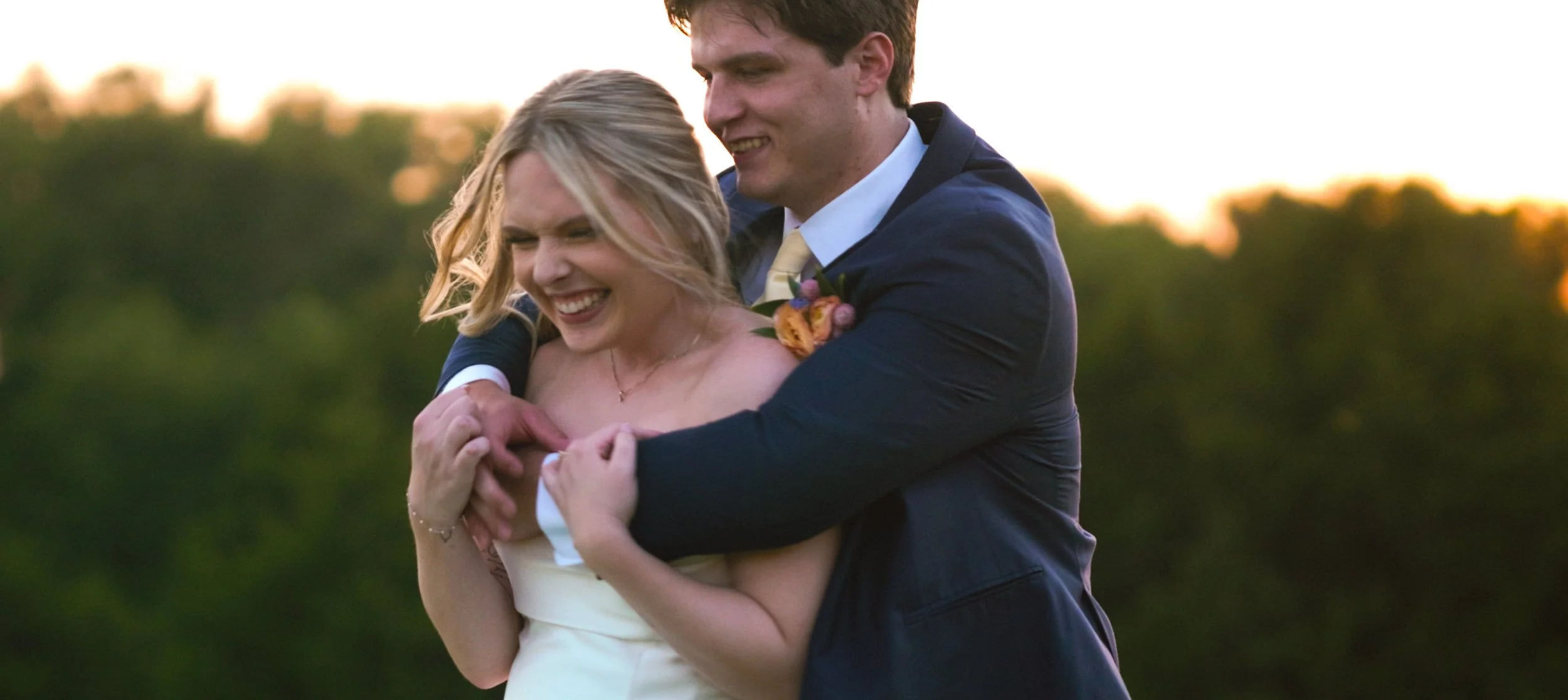 A joyful couple dressed in wedding attire hugging outdoors during sunset with trees in the background.