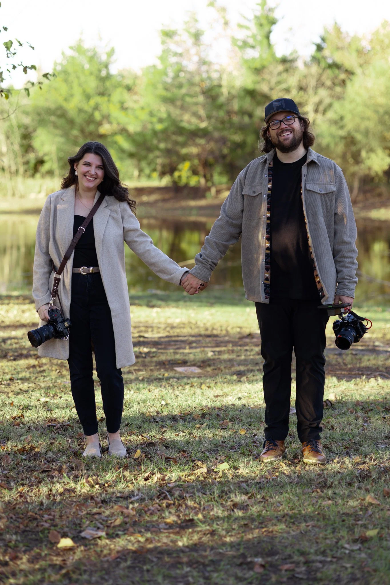 A smiling couple holding hands outdoors by a lake in a park, both holding cameras, with lush trees in the background.
