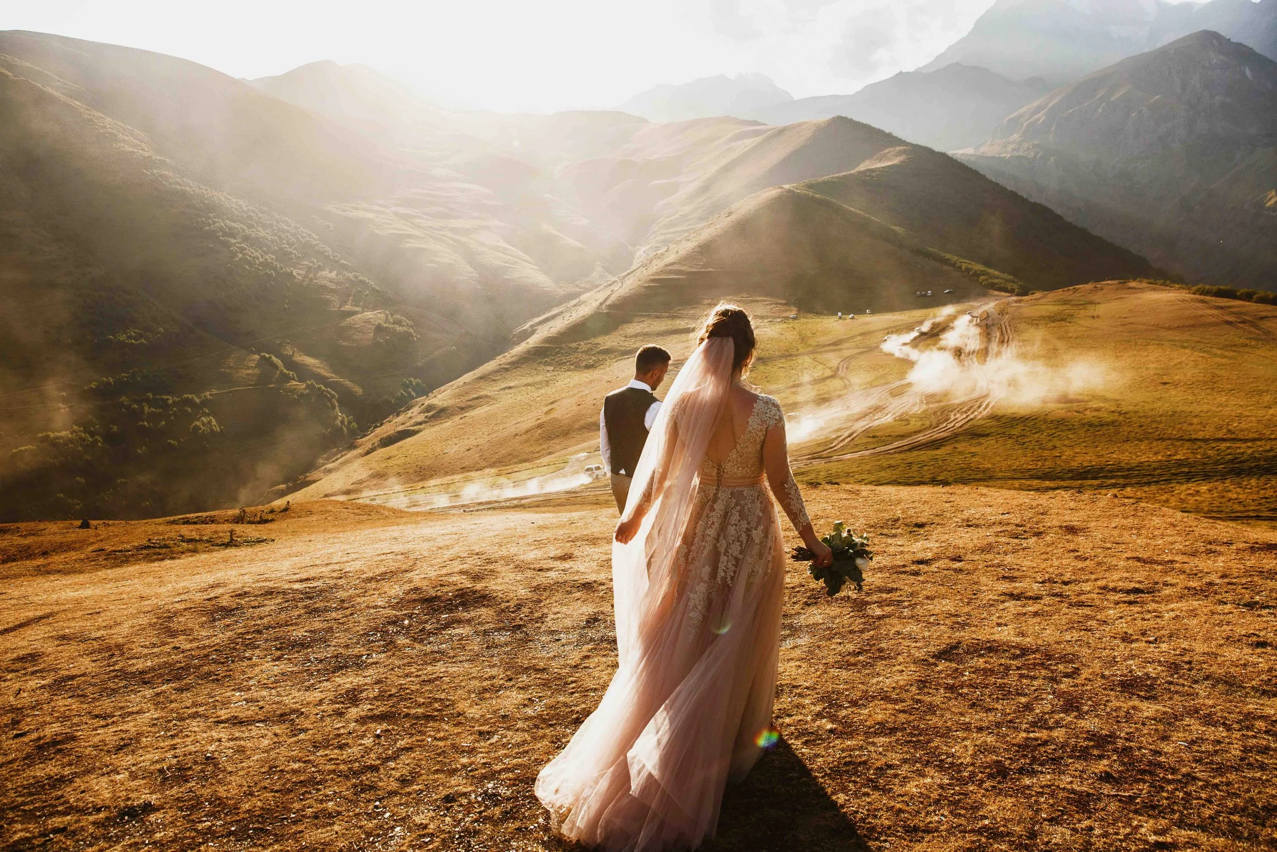 A bride and groom walking in a mountainous landscape with steam rising from the ground during sunset.