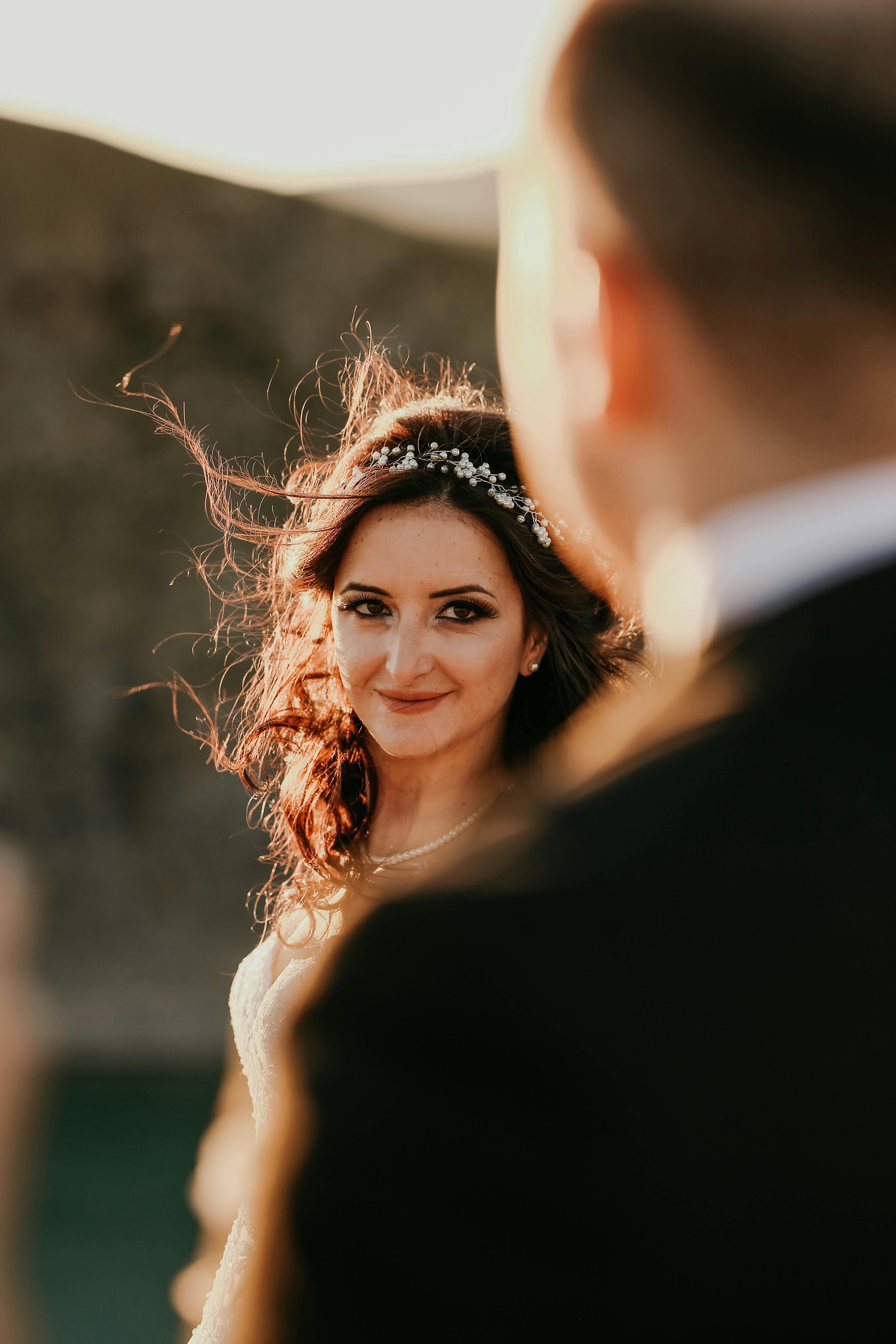 Close-up of a bride with long, wavy hair wearing a pearl necklace and headband, smiling at the camera, with a groom partially visible in the foreground.