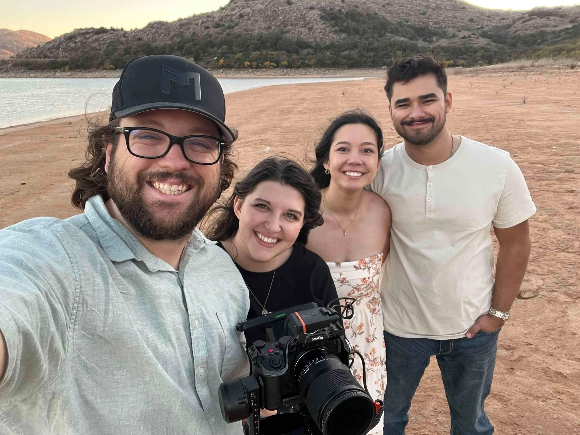 Group of four friends smiling at the camera on a sandy lakeside with mountains in the background.