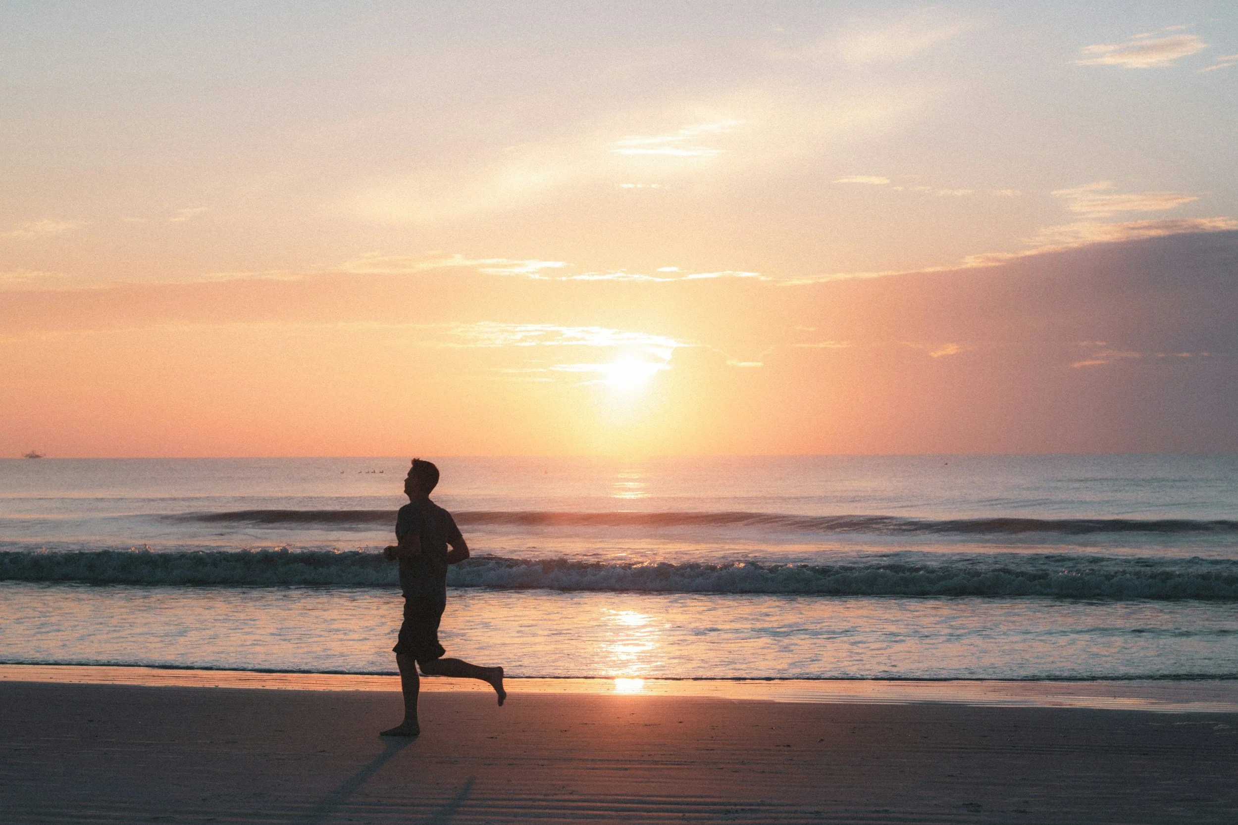 A person running on the beach during a sunset, with the ocean in the background and the sky painted with soft pastel colors.