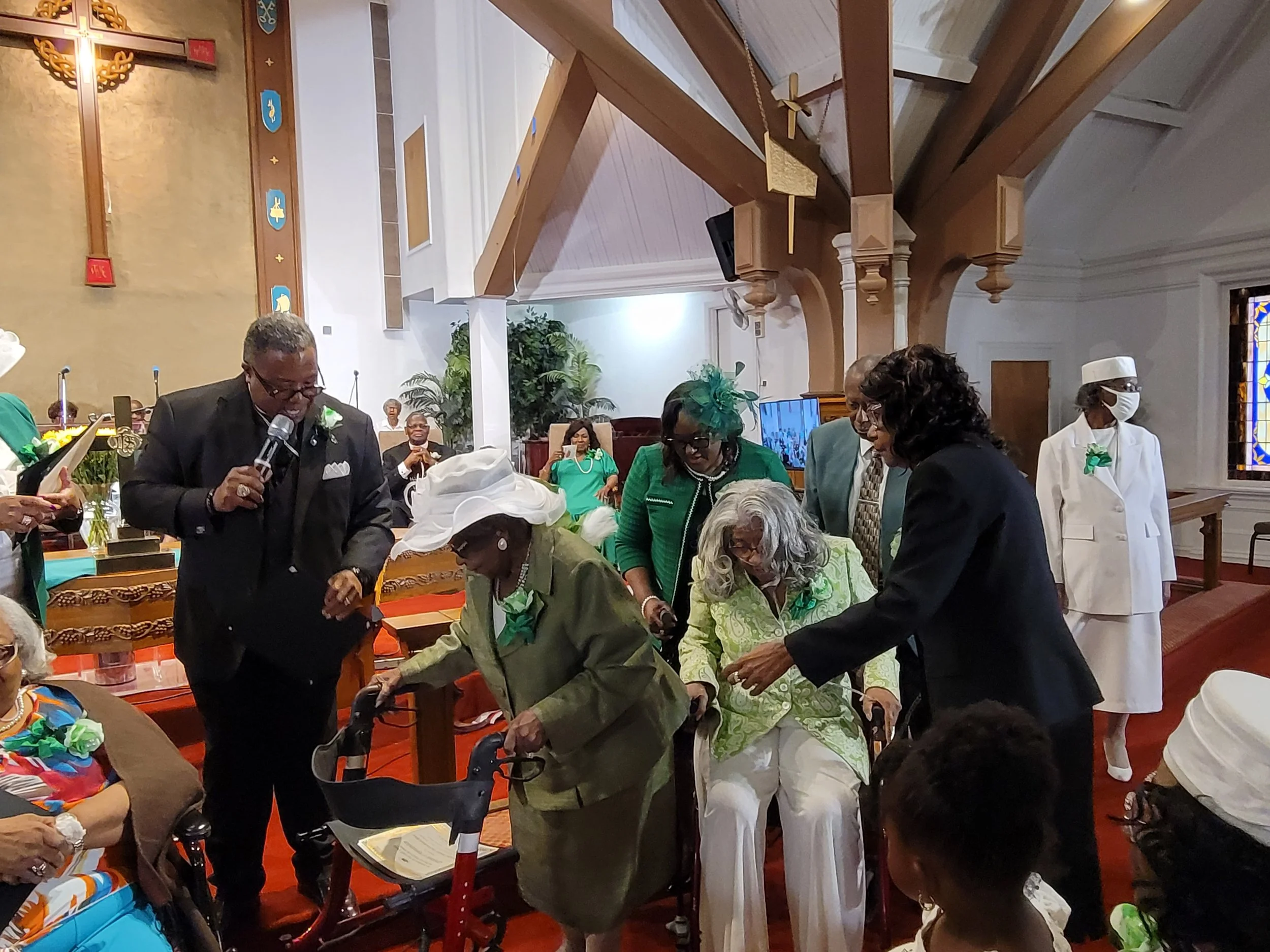 Group of elderly women and men, some using walkers, gathered inside a church for a celebration. One woman is being assisted in her walker by a person in a black suit. Several people are dressed in green, with some wearing hats and corsages. The churc
