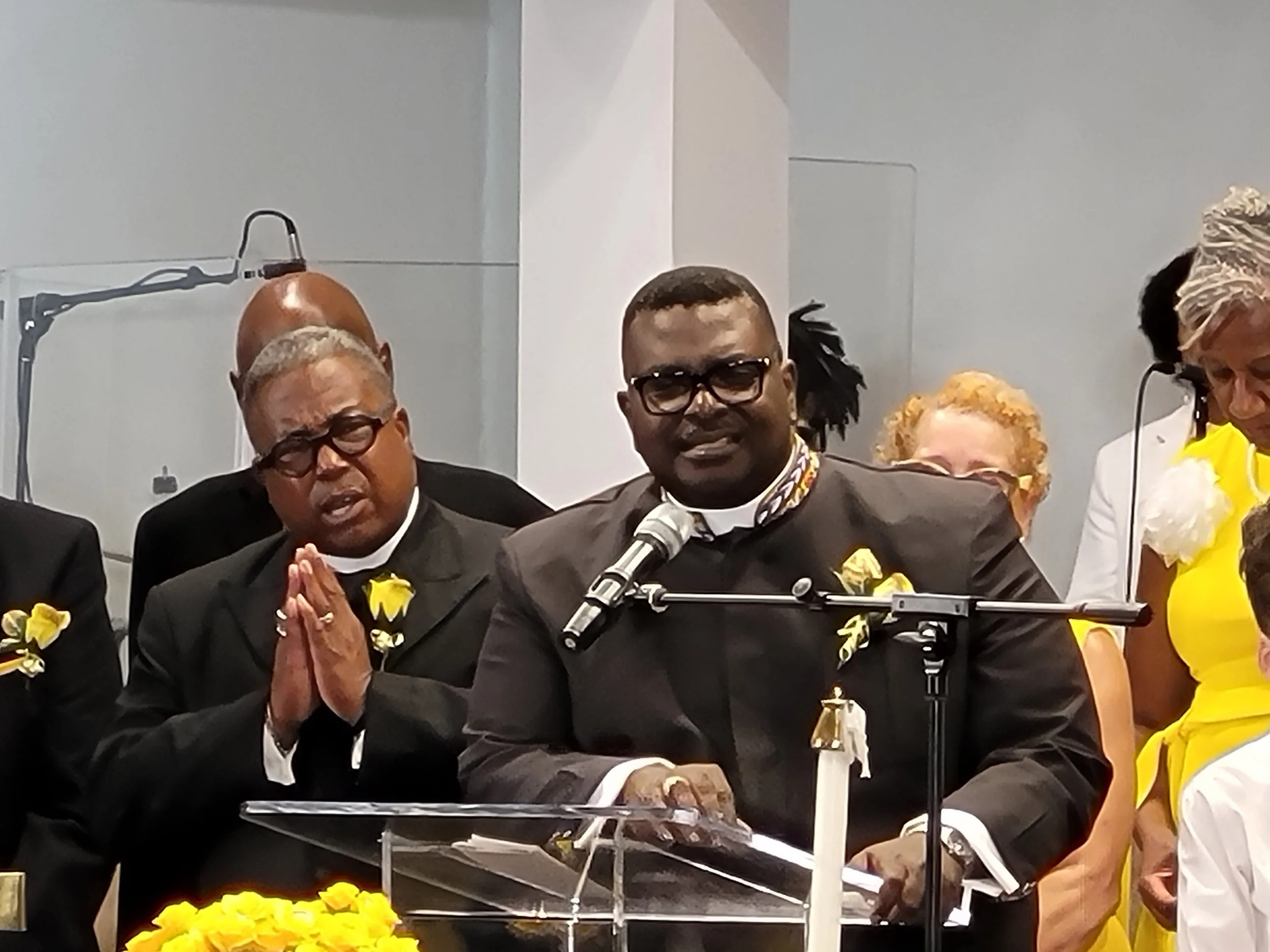 A man dressed in a black suit and glasses speaking at a podium with a microphone, surrounded by people in formal attire at a formal event, with some people praying or listening attentively.