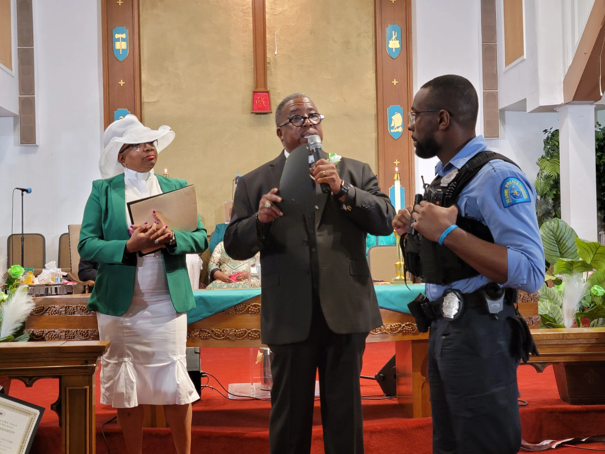 A woman in a white hat and green blazer holding a folder, a man in a dark suit speaking into a microphone, and a police officer in uniform standing next to the speaker, inside a church or chapel with religious symbols on the wall.