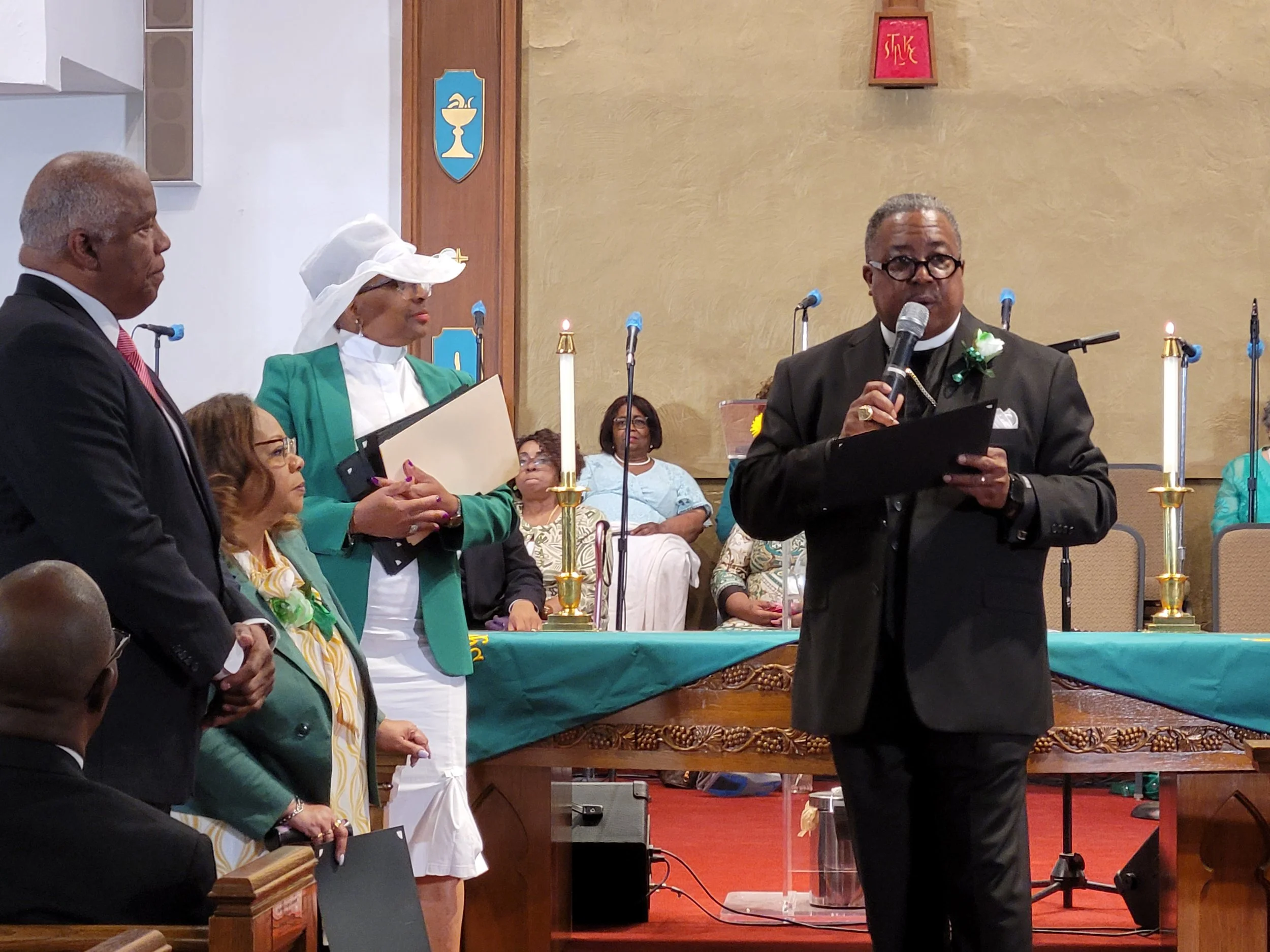 A man in a black suit and glasses speaking into a microphone at a church service or ceremony, with a group of people sitting or standing nearby and candles on the altar.