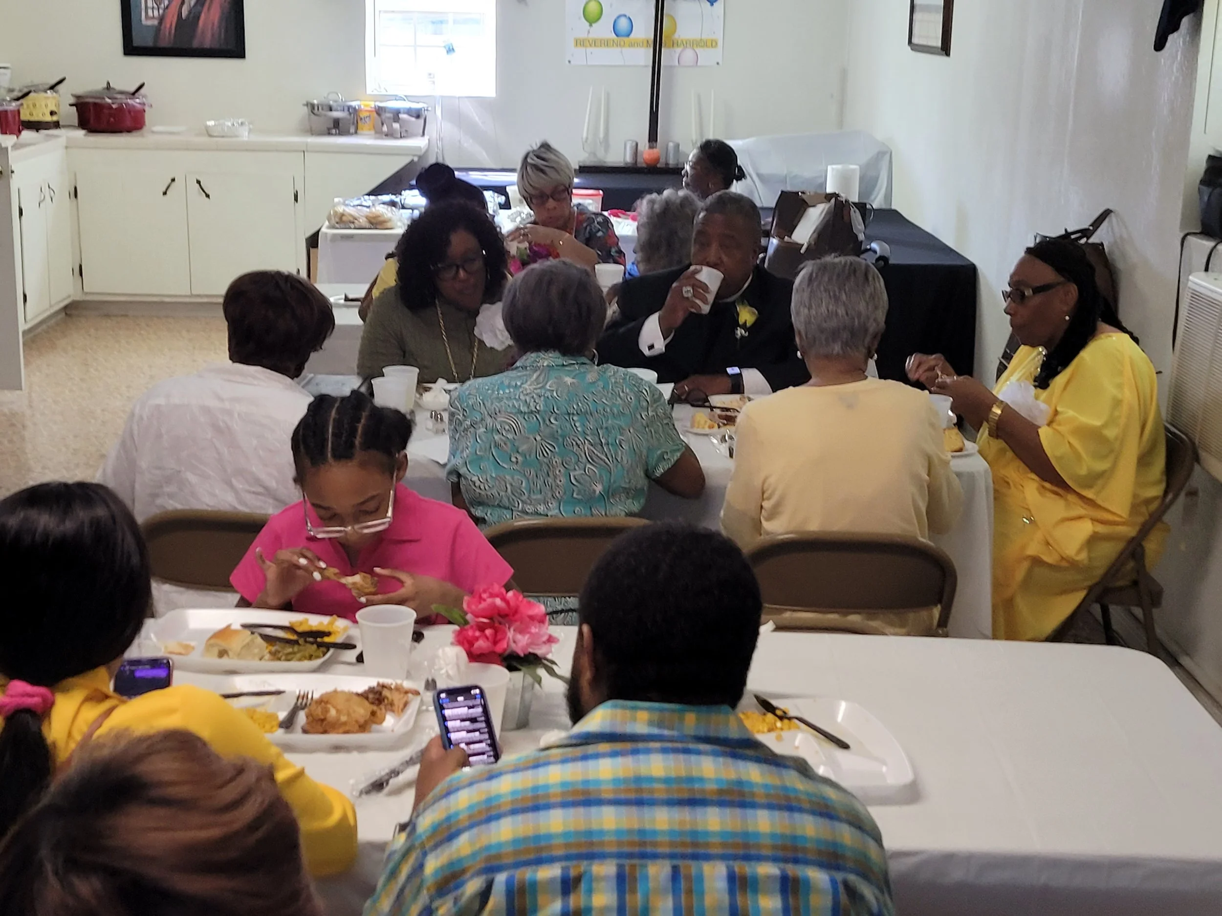 Group of people, mostly older adults, sitting around a table at a social gathering, eating and conversing. A young girl in pink is eating, and some people are using their phones. The setting appears to be a community hall or similar venue.