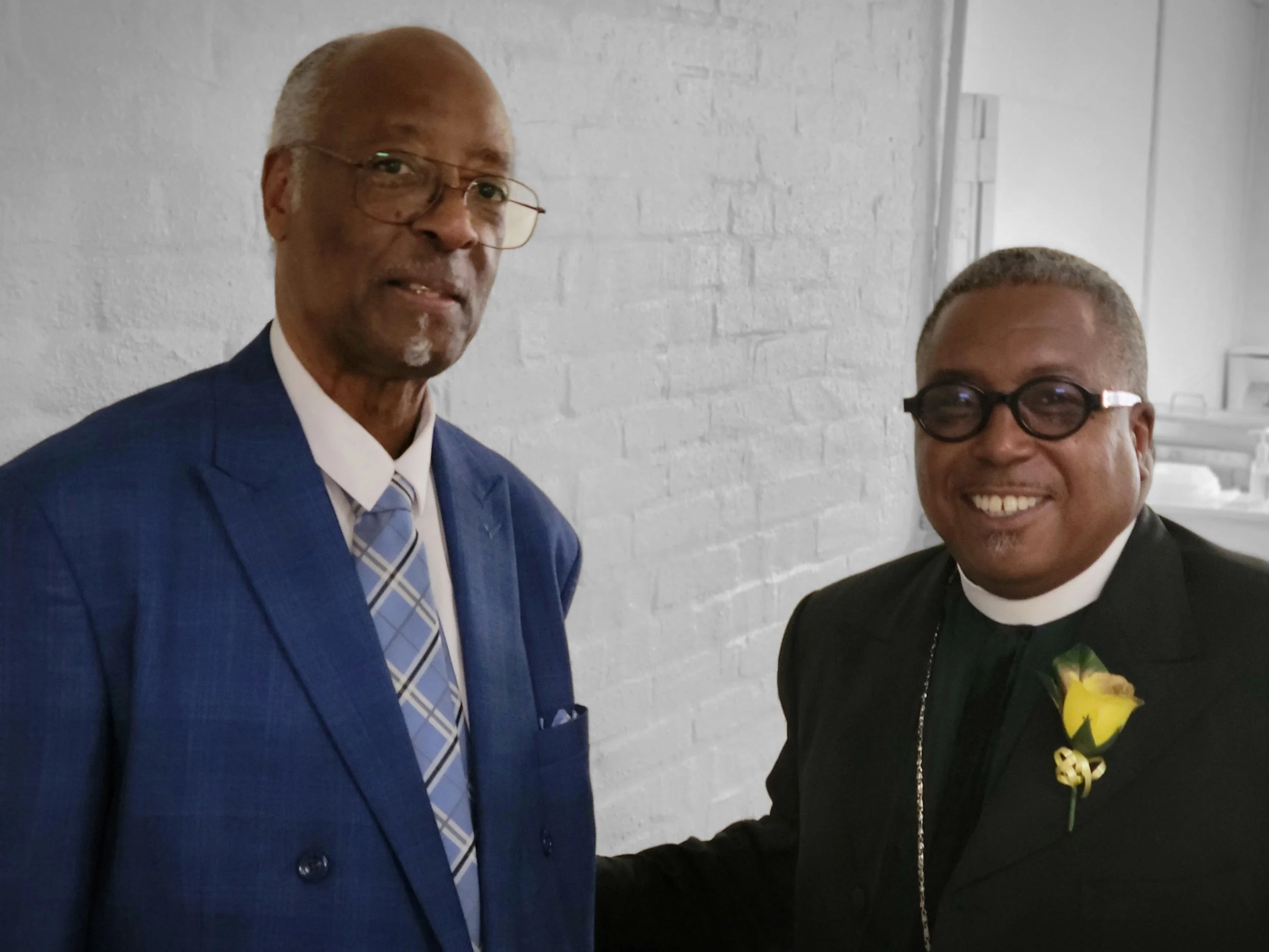 Two men dressed in formal attire standing next to each other, smiling, in front of a white brick wall.
