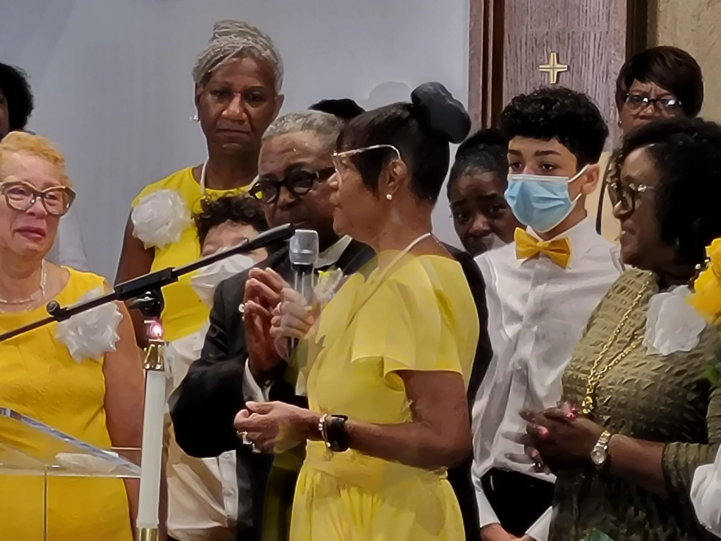A group of people gathered at a church service, with some wearing yellow attire and floral decorations, including a woman speaking into a microphone, a woman with glasses and a black dress, and a young man wearing a face mask and yellow bow tie.