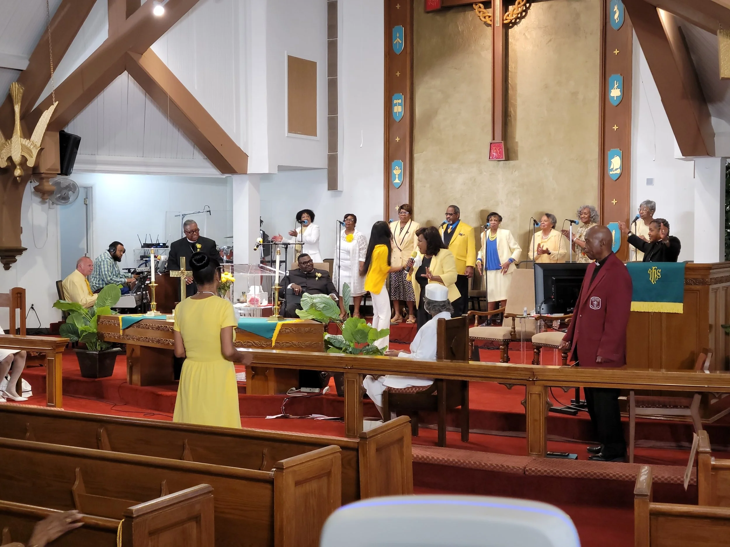 A church service with a choir singing on stage, a person speaking at the front, and congregation members seated and standing in the pews.