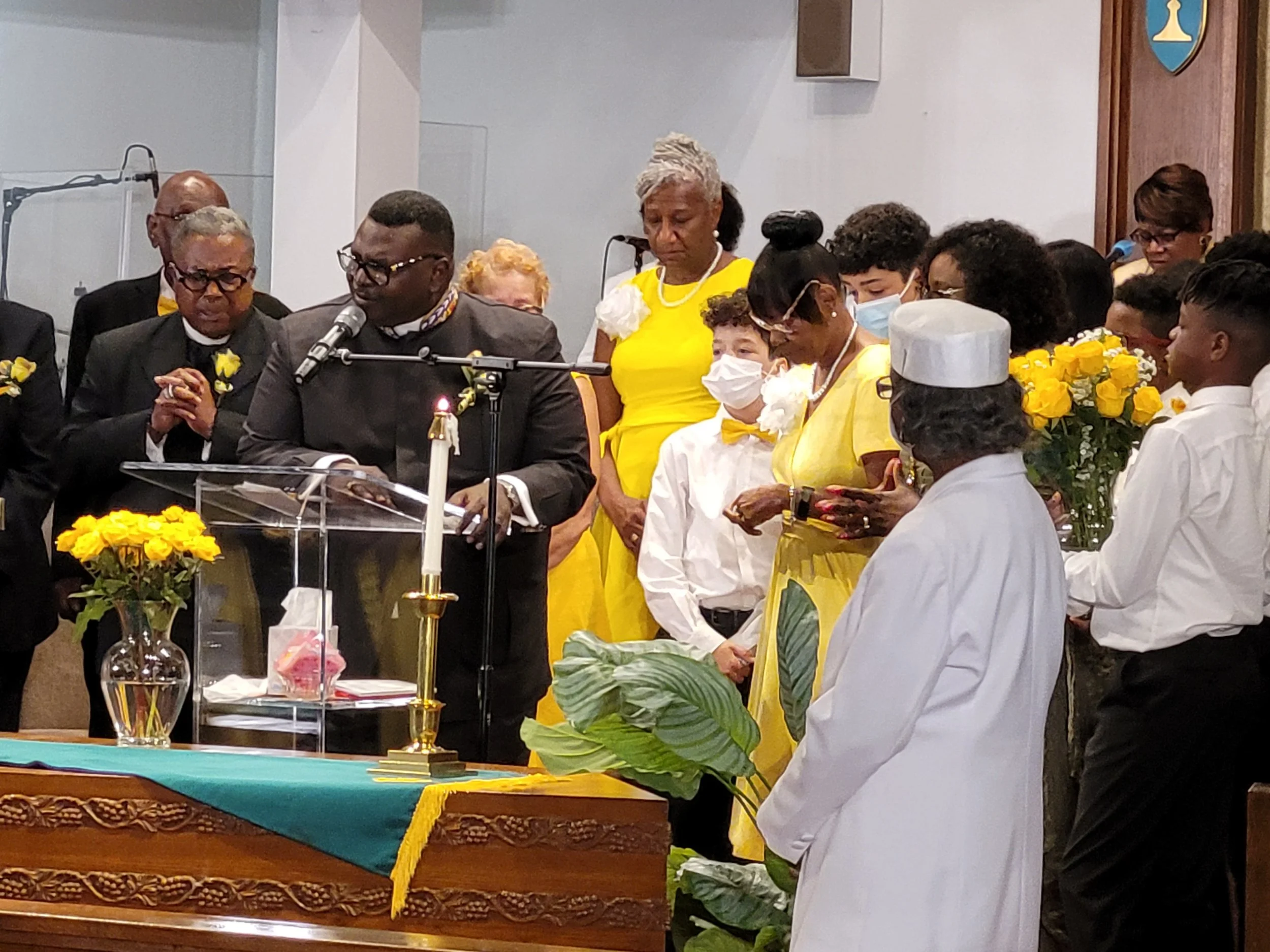 A diverse group of people at a church service, with some praying, holding flowers, or reading from a book, behind a table with flowers, candle, and religious items, in a church decorated with a shield-shaped emblem.