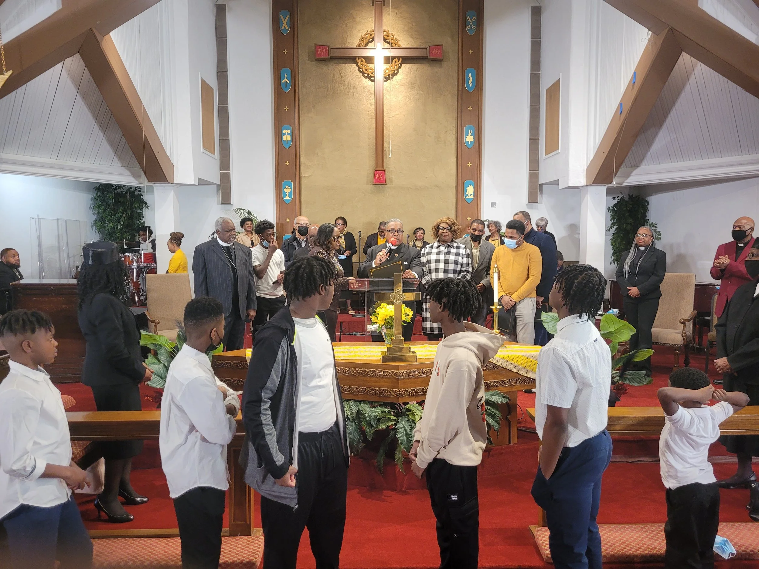A religious ceremony taking place in a church with a cross at the back wall, and a congregation facing forward. Children are standing in front of the altar, and adults are gathered behind them, some wearing masks.