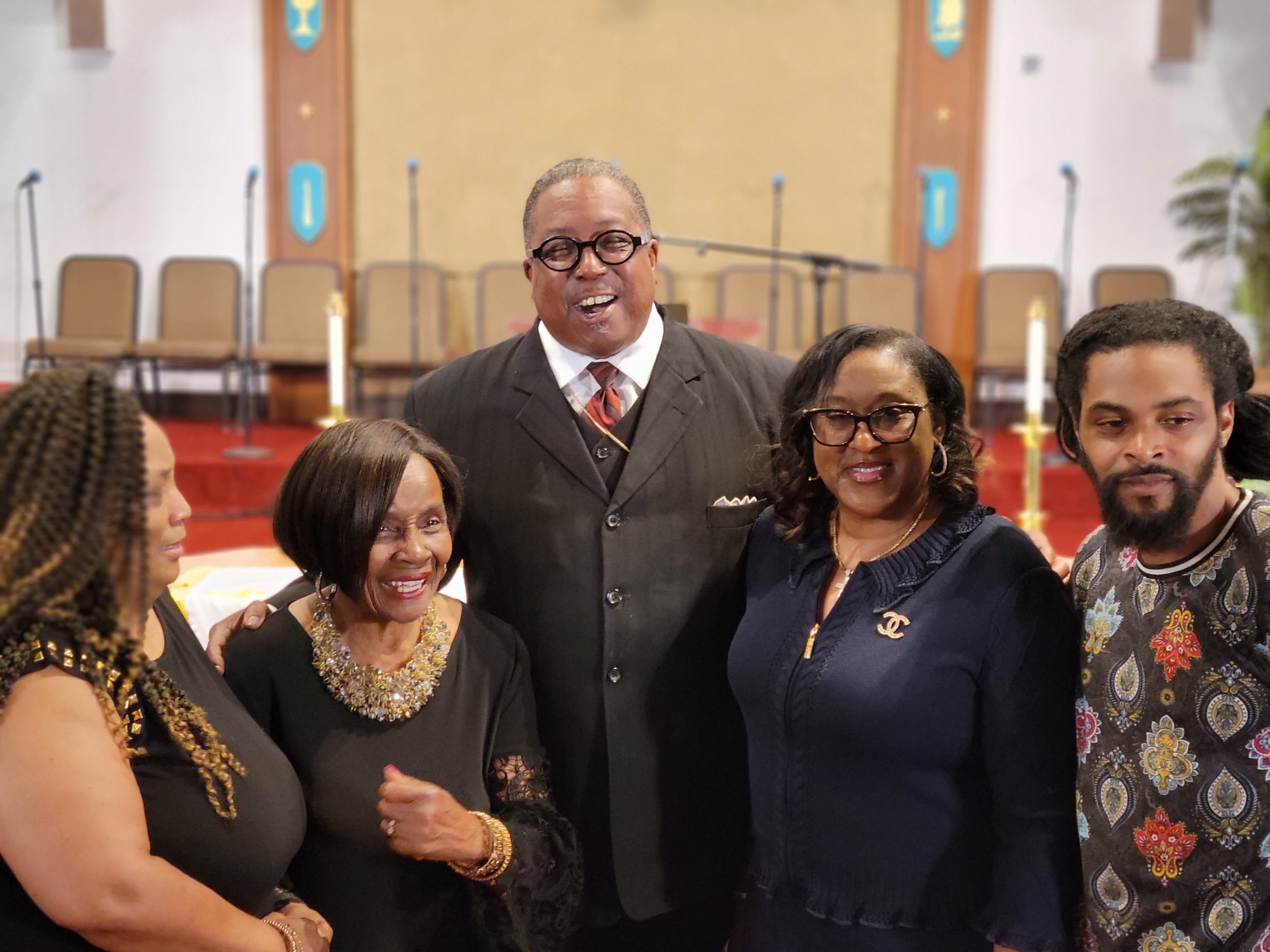 Group of five African American people, three women and two men, smiling and posing together in a church or event space with a stage and chairs in the background.