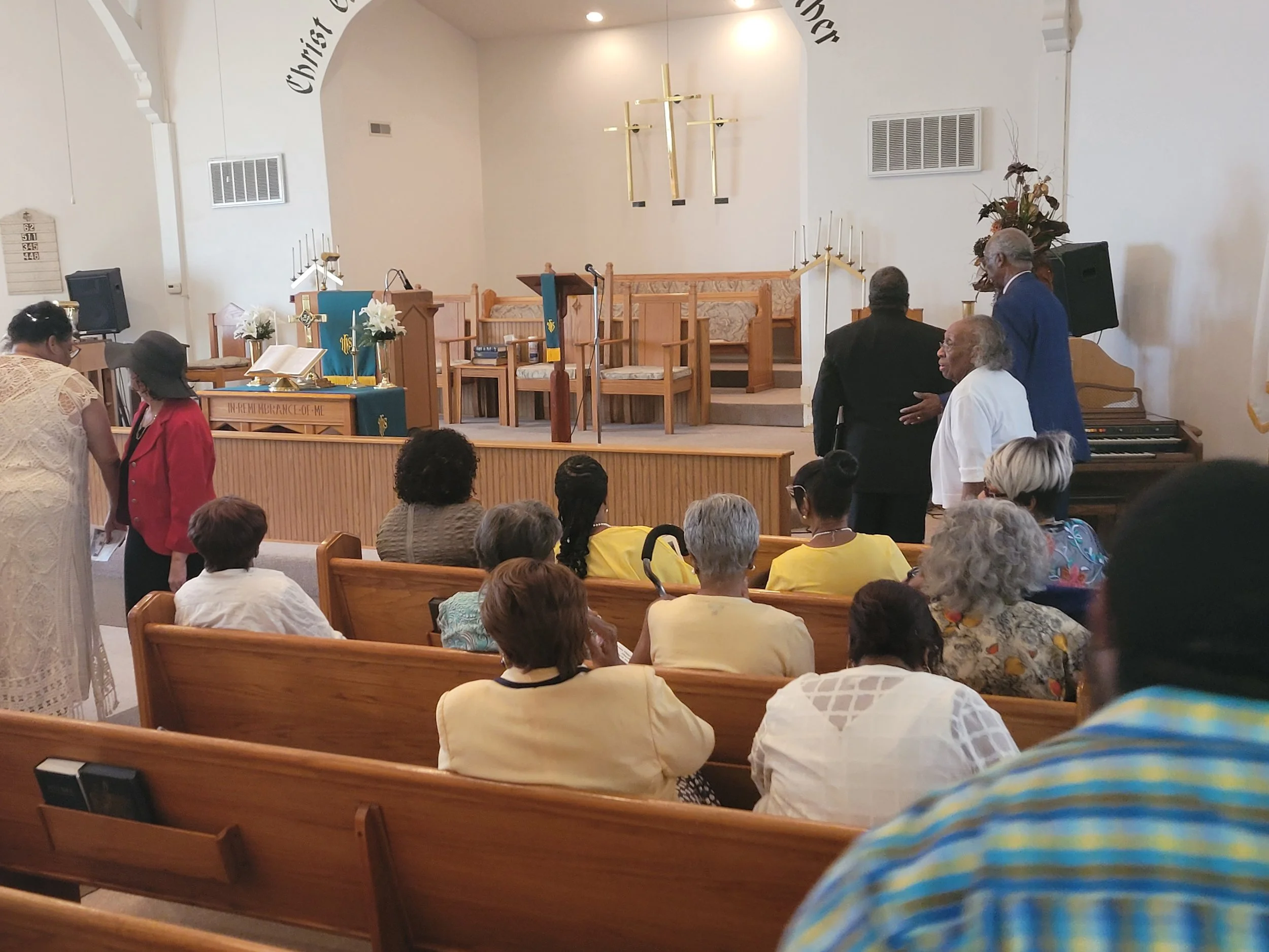 A diverse group of church members gathered inside a church with wooden pews, preparing for a service or event. The front stage area includes an altar with Christian symbols, flowers, and a Bible, with a microphone and musical keyboard nearby.