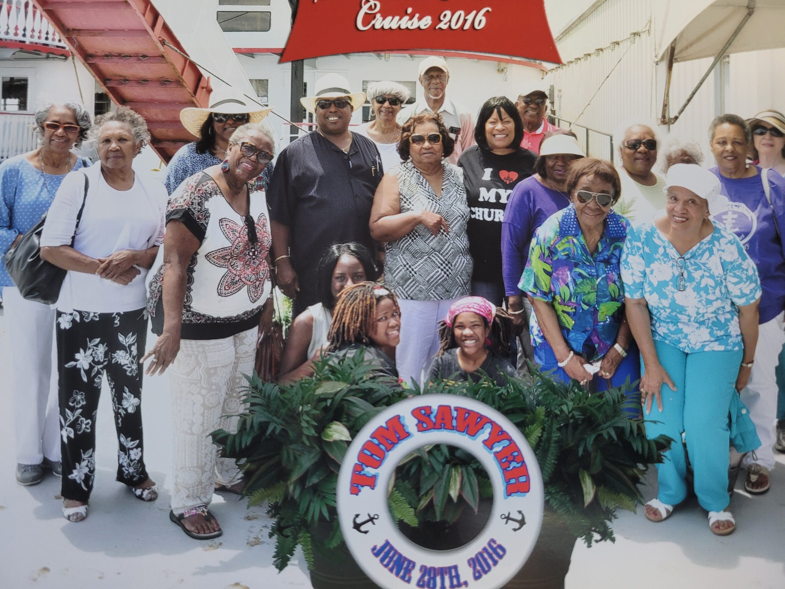 Group of people posing at a cruise event in June 2016, with a sign that reads 'Tom Sawyer Cruise 2016' and a floral arrangement in front of them.