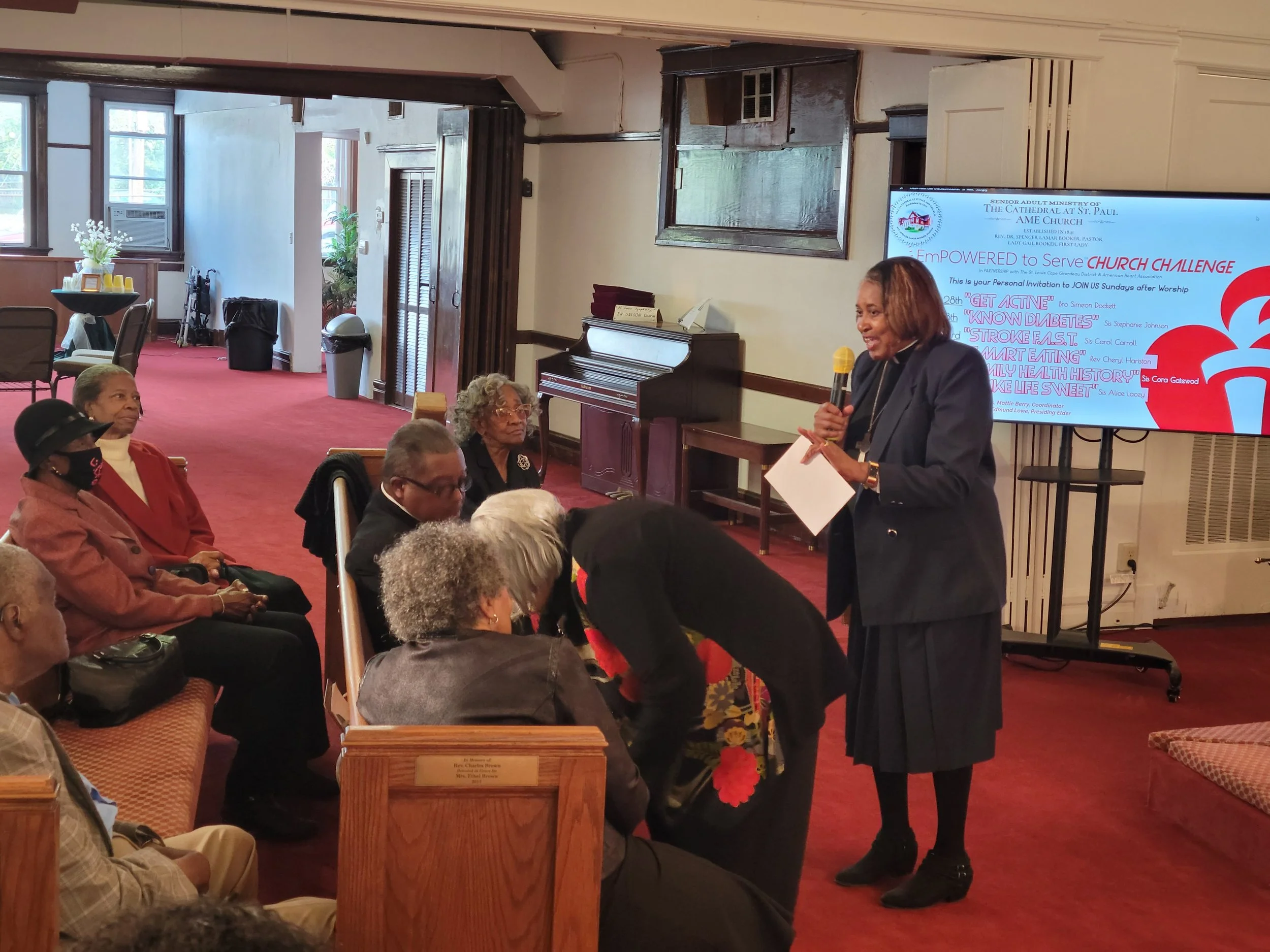 A woman giving a presentation to an audience in a church or community room. She is holding a microphone and a paper, standing in front of a large screen with event details. The audience is seated, with some taking notes or listening attentively.