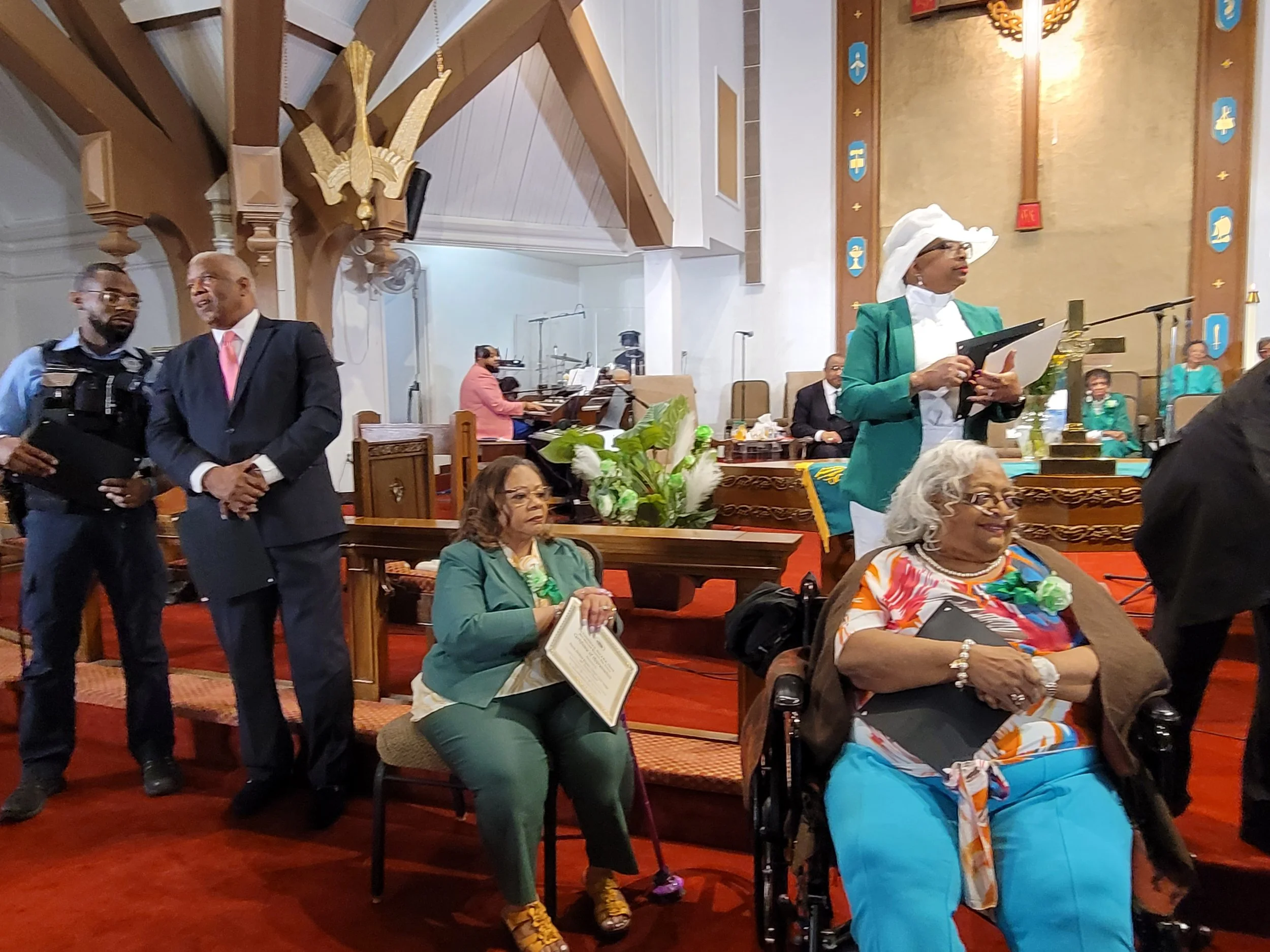 A church service or event with a diverse group of people, including women, seated and standing in a church interior with wood accents and shields on the wall. One woman is standing and reading, while two women are seated in the foreground, one holdin