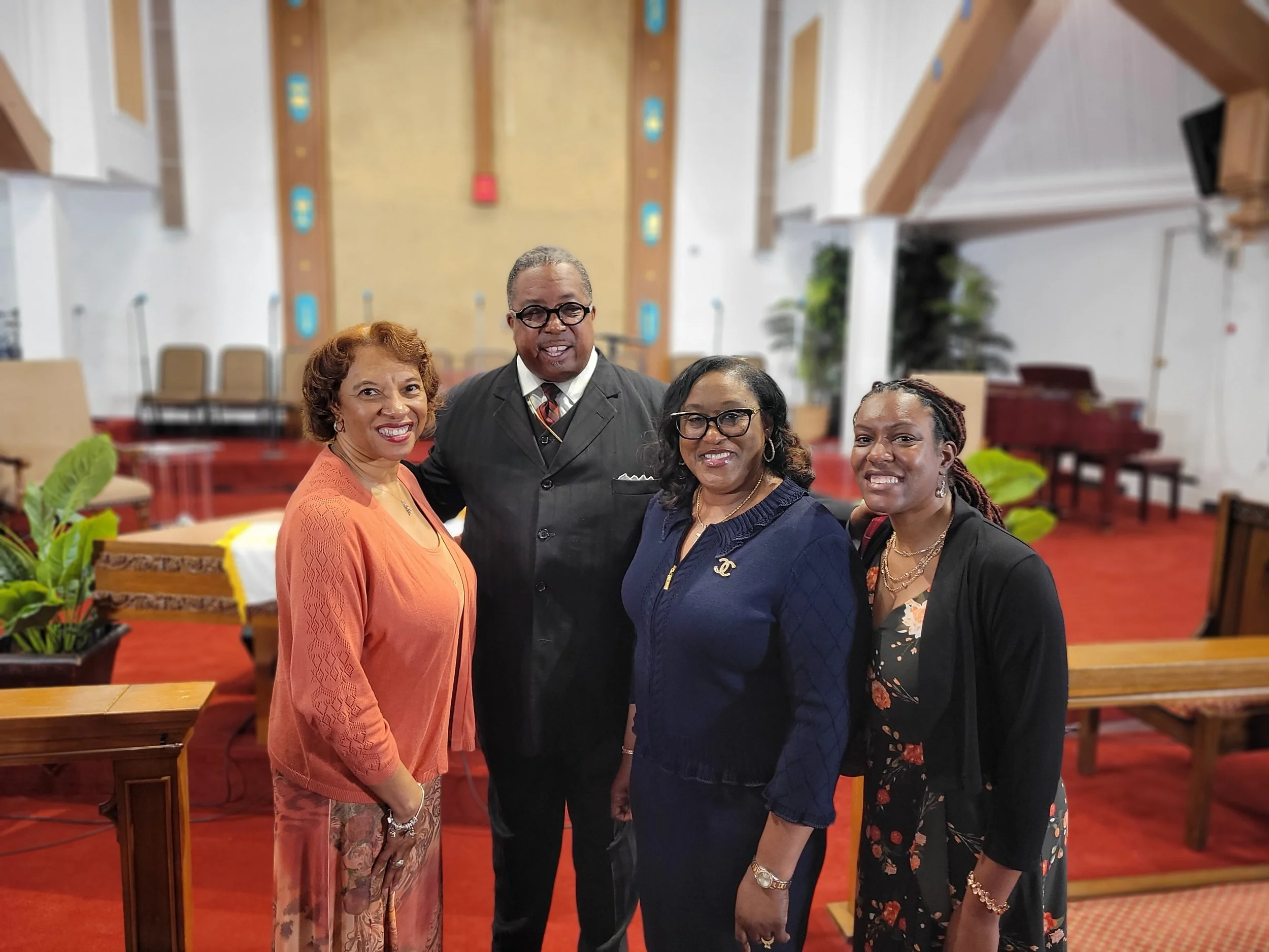 Four people, three women and one man, are standing together inside a church. They are smiling and dressed in formal attire, with the women wearing dresses or suits and the man in a suit and tie. The background shows a stage with chairs and musical in