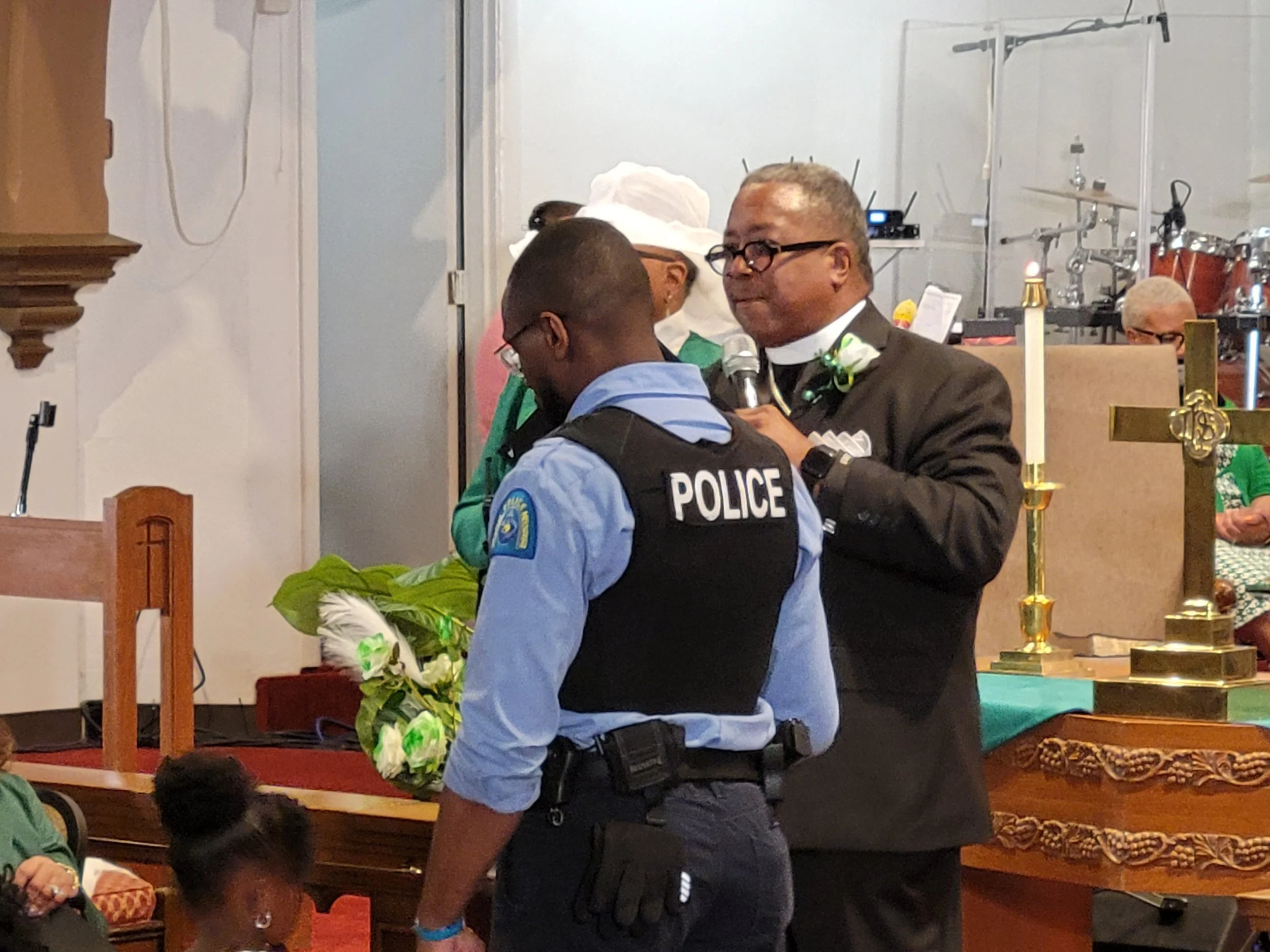 A police officer and a clergyman at a ceremony inside a church, with floral arrangements and religious artifacts.