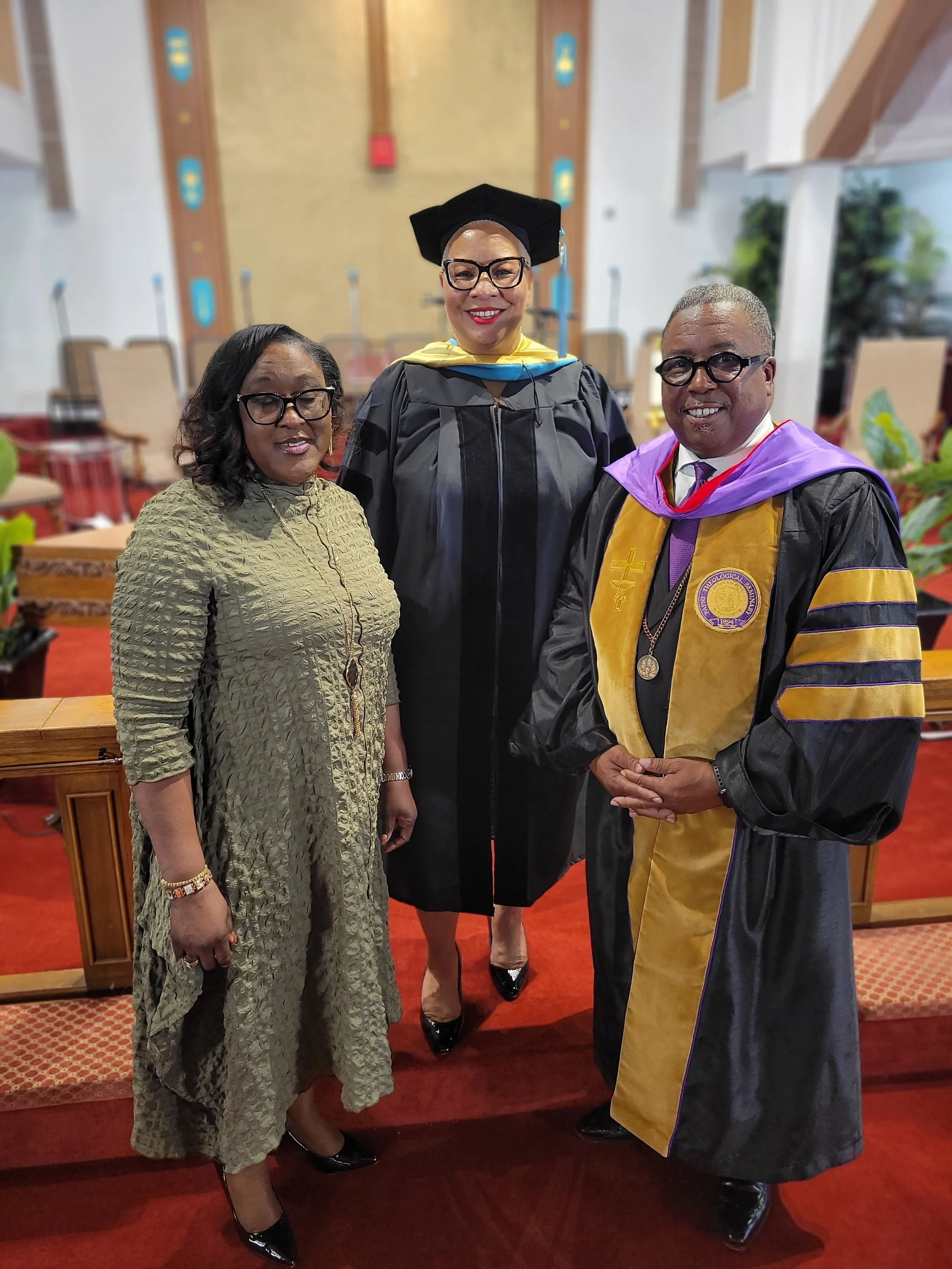 A woman in a green dress, a woman in a graduation gown and cap, and a man in an academic gown and hood standing together inside a room with wooden furniture and red carpet.