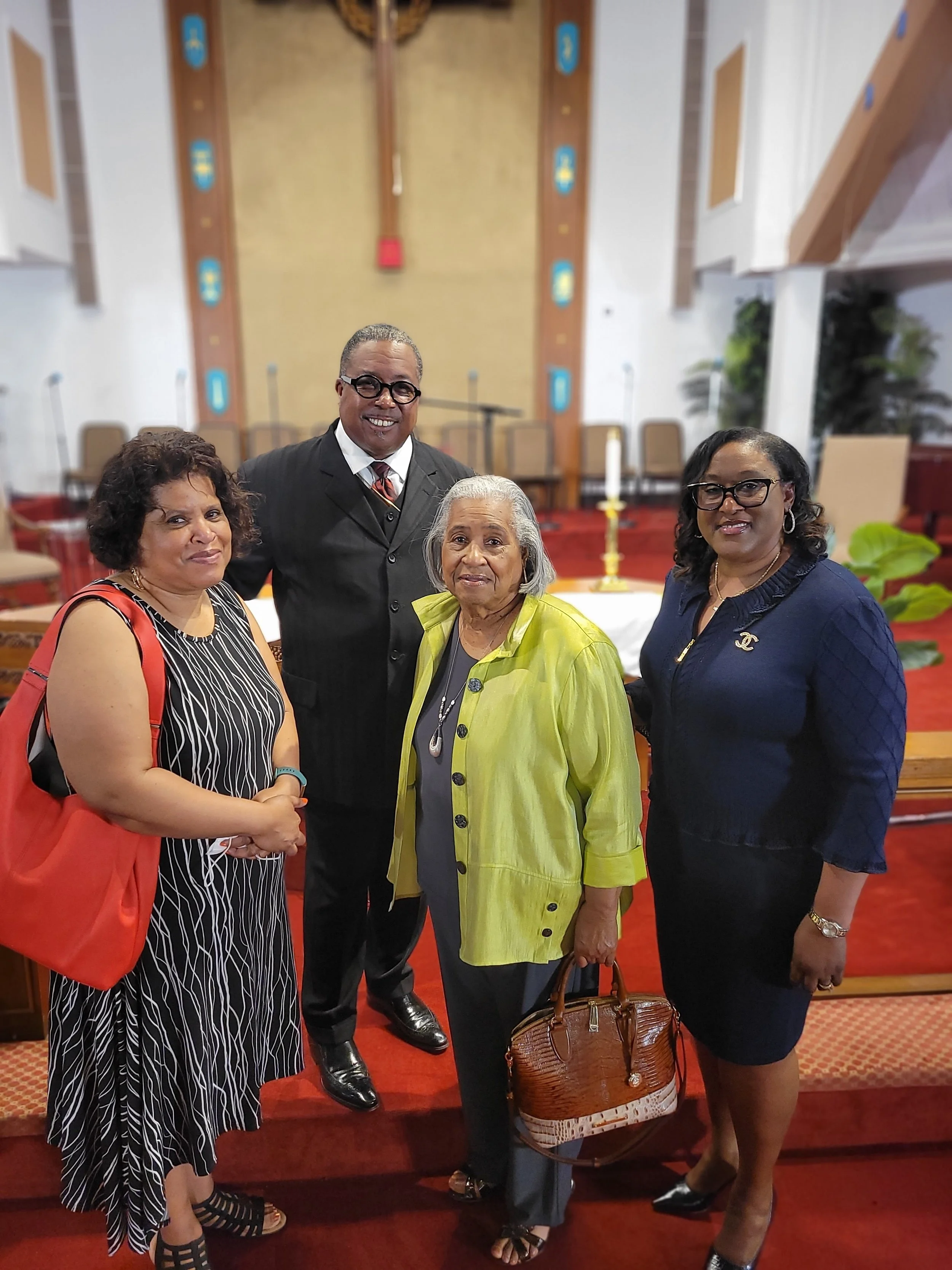 Four women and one man standing together inside a church with a large cross on the wall behind them. The women are dressed in colorful clothing, and the man is wearing a suit and tie. They are smiling at the camera.