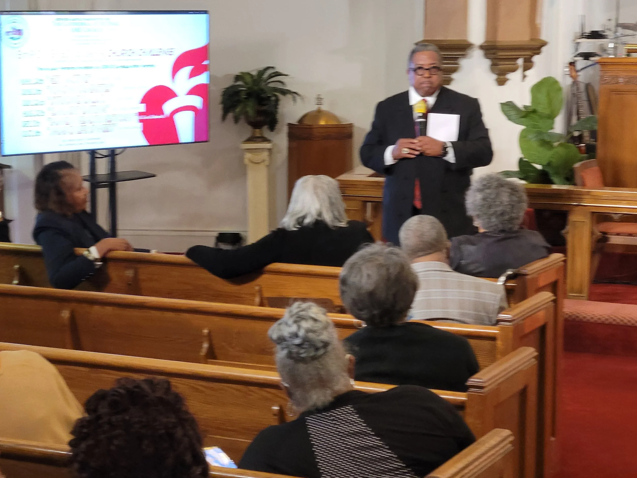 A man in a suit speaking at a presentation in a church-like setting with an audience seated on wooden pews. There is a large screen displaying a logo and text behind him.