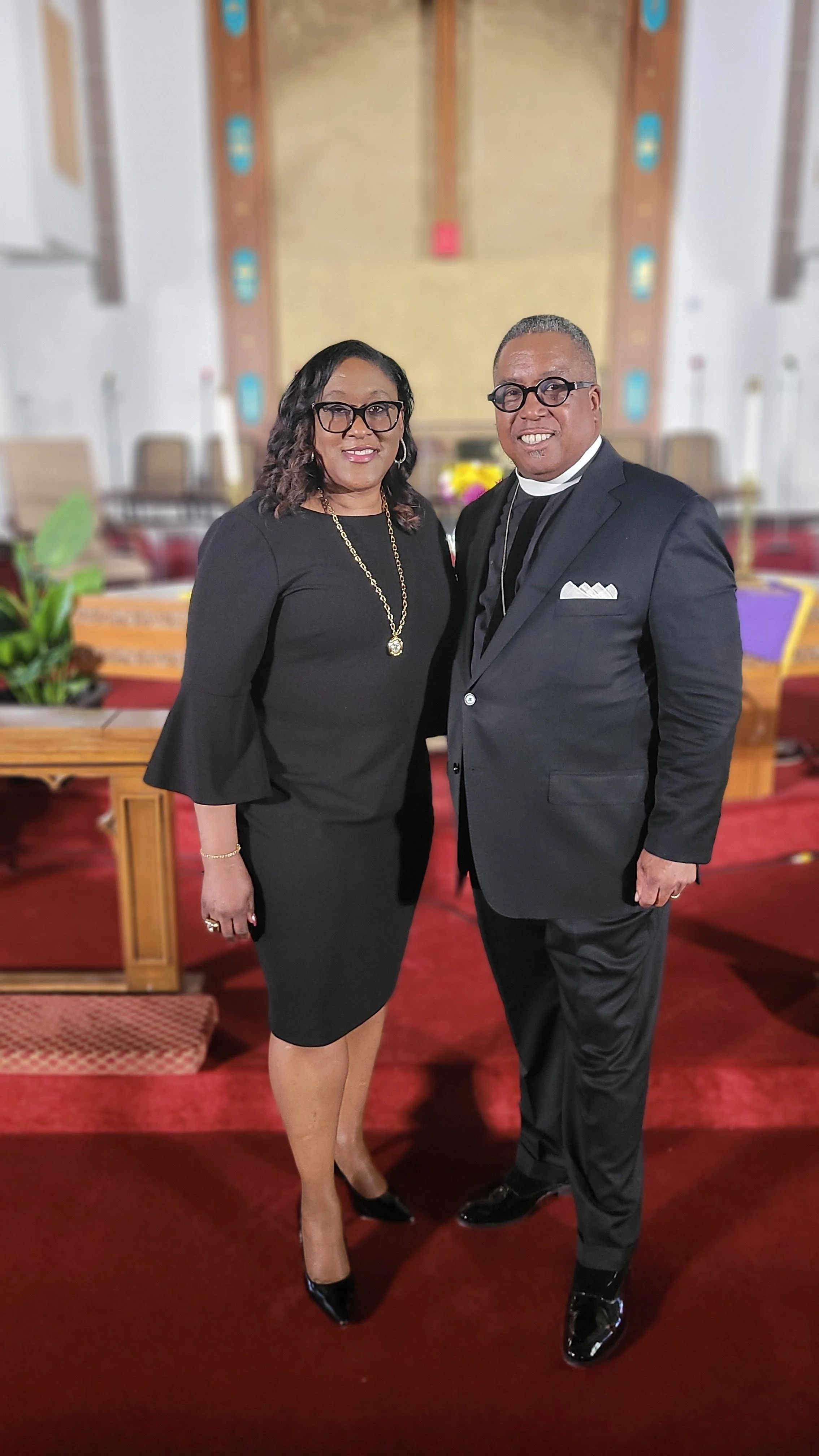 A woman and a man standing together in a church, dressed in formal black attire. The woman wears a black dress with bell sleeves, black heels, and jewelry. The man wears a black suit, clerical collar, and glasses.