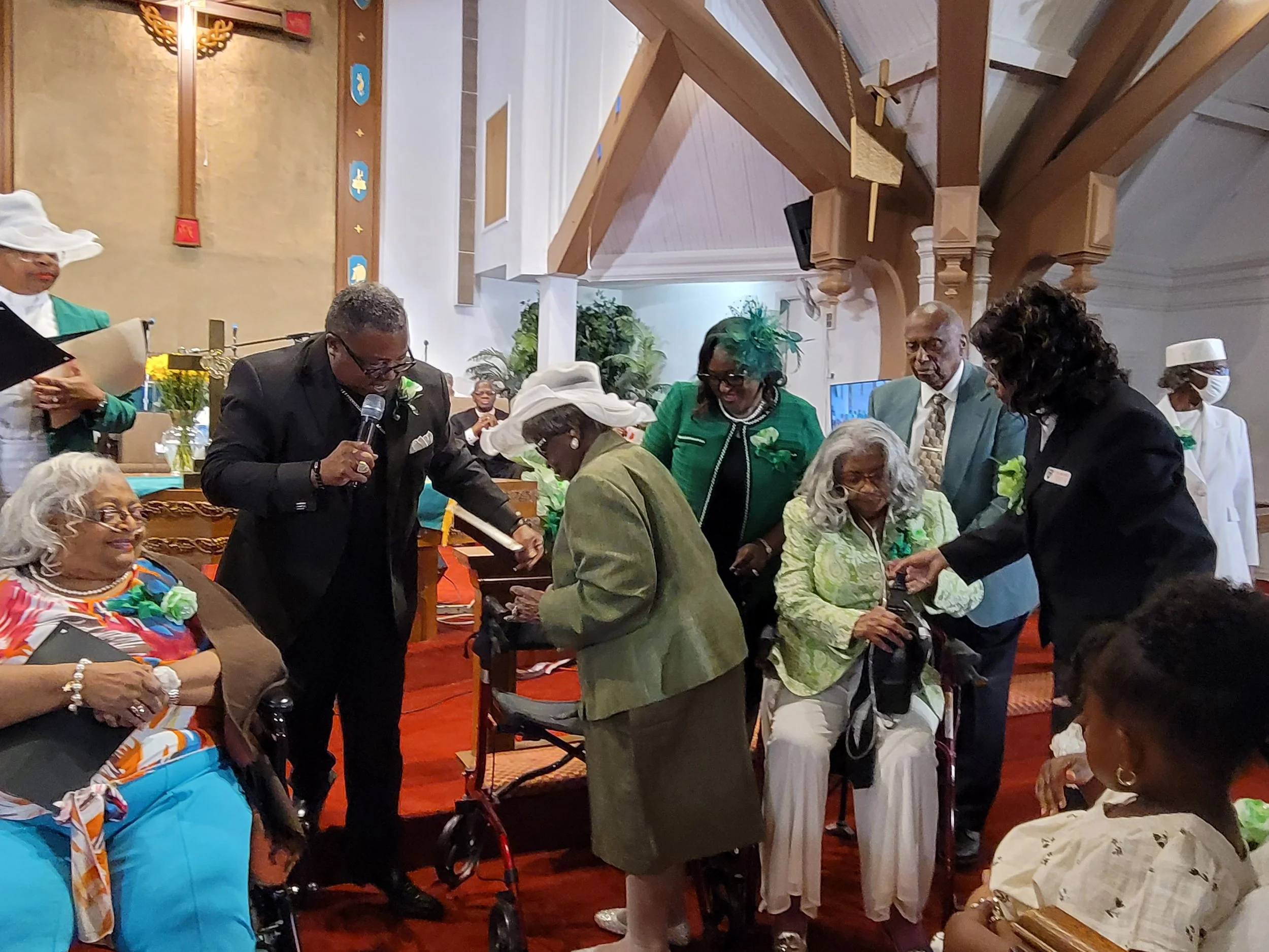 Group of elderly women and men dressed in colorful and formal attire gathered inside a church, engaging in a celebration or event, with some older women seated and others standing, some using a walker, and a man holding a microphone.