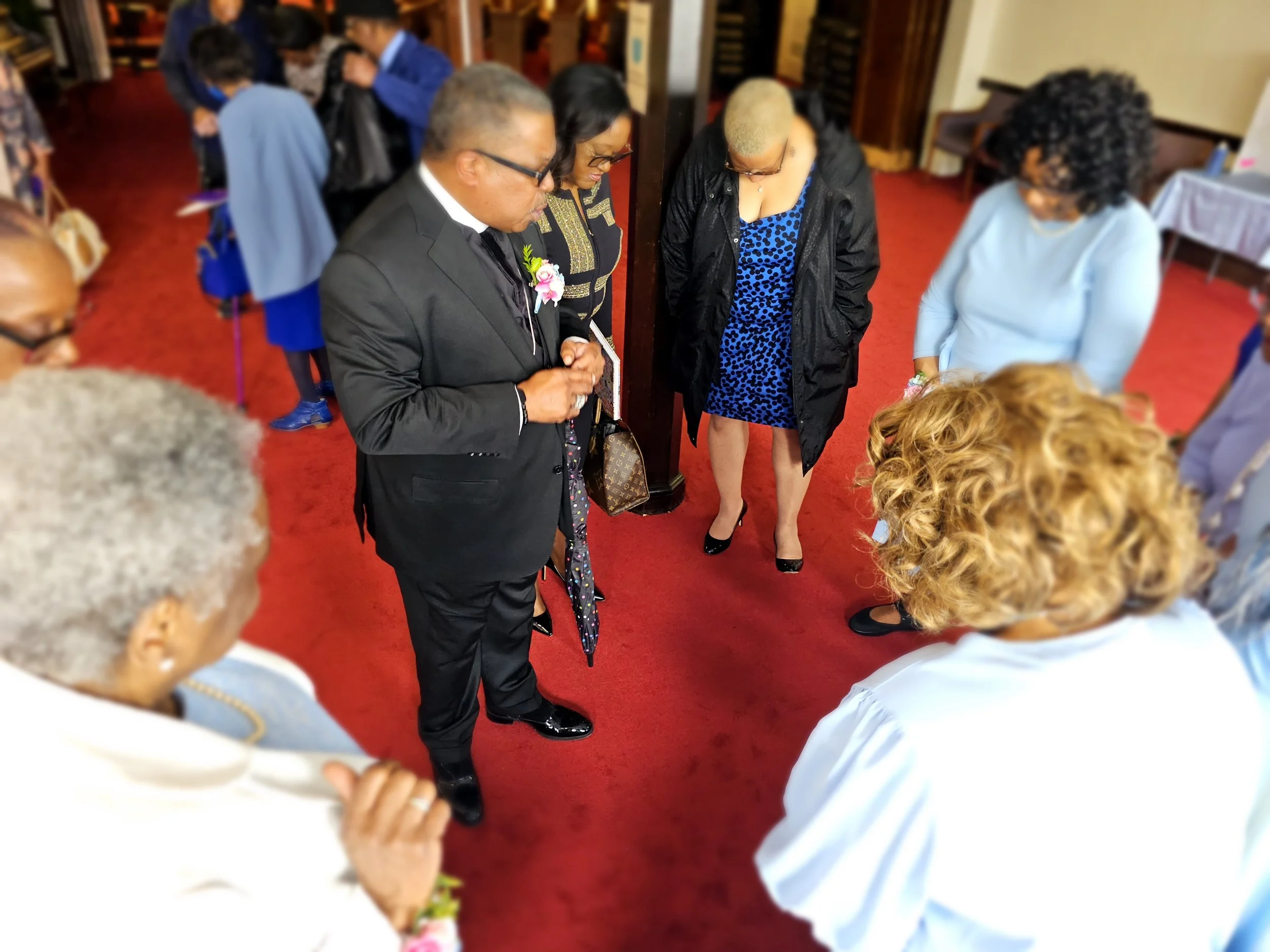 A group of people are gathered in a prayer circle, heads bowed, with some holding hands or clasping their hands in front of them, in what appears to be a religious or solemn gathering inside a building with a red carpeted floor.