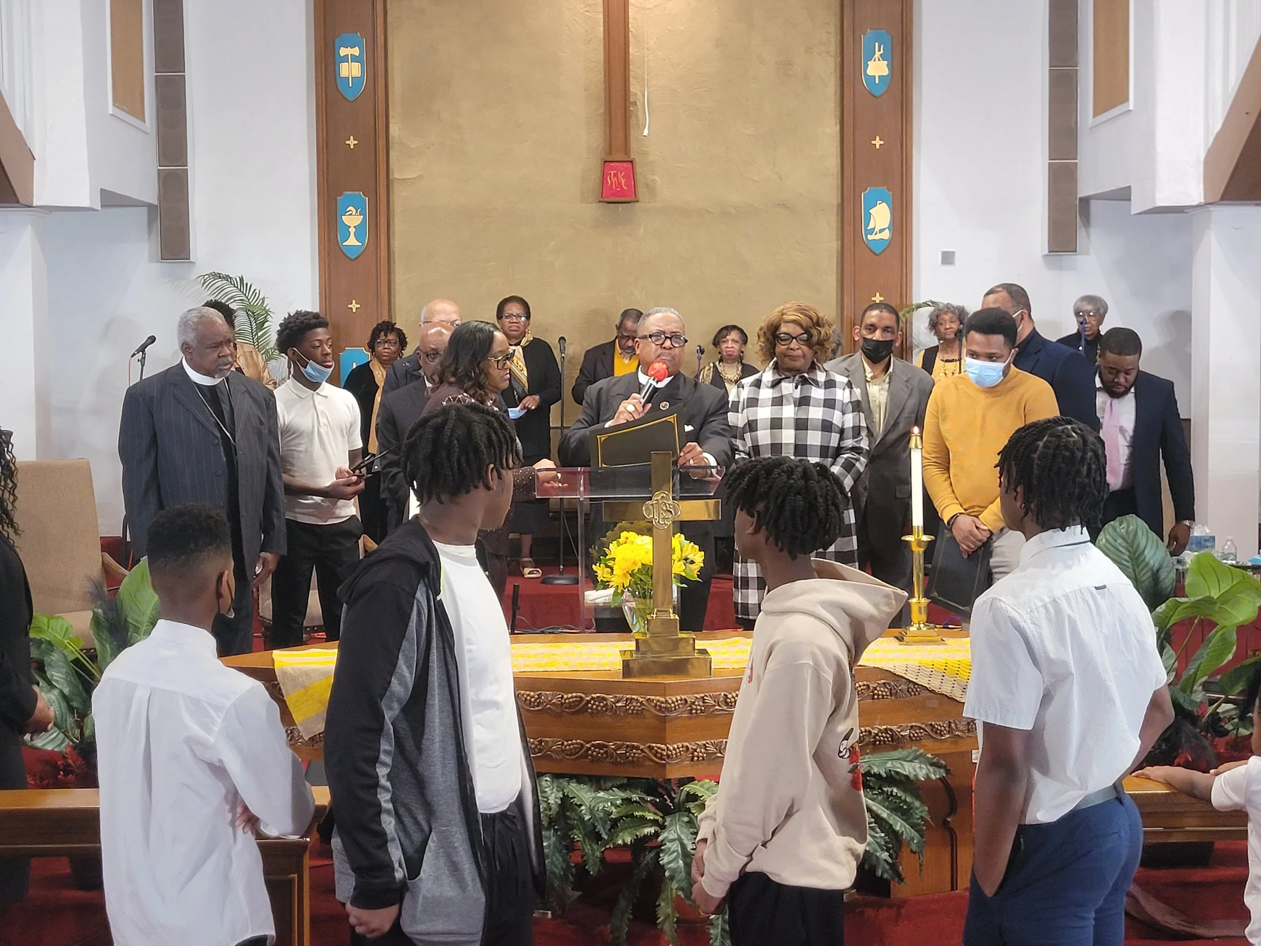 A group of people gathered in a church, with some standing behind the altar and others facing each other in front. The altar has a yellow cloth, a candle, and flowers. Several individuals are wearing face masks.