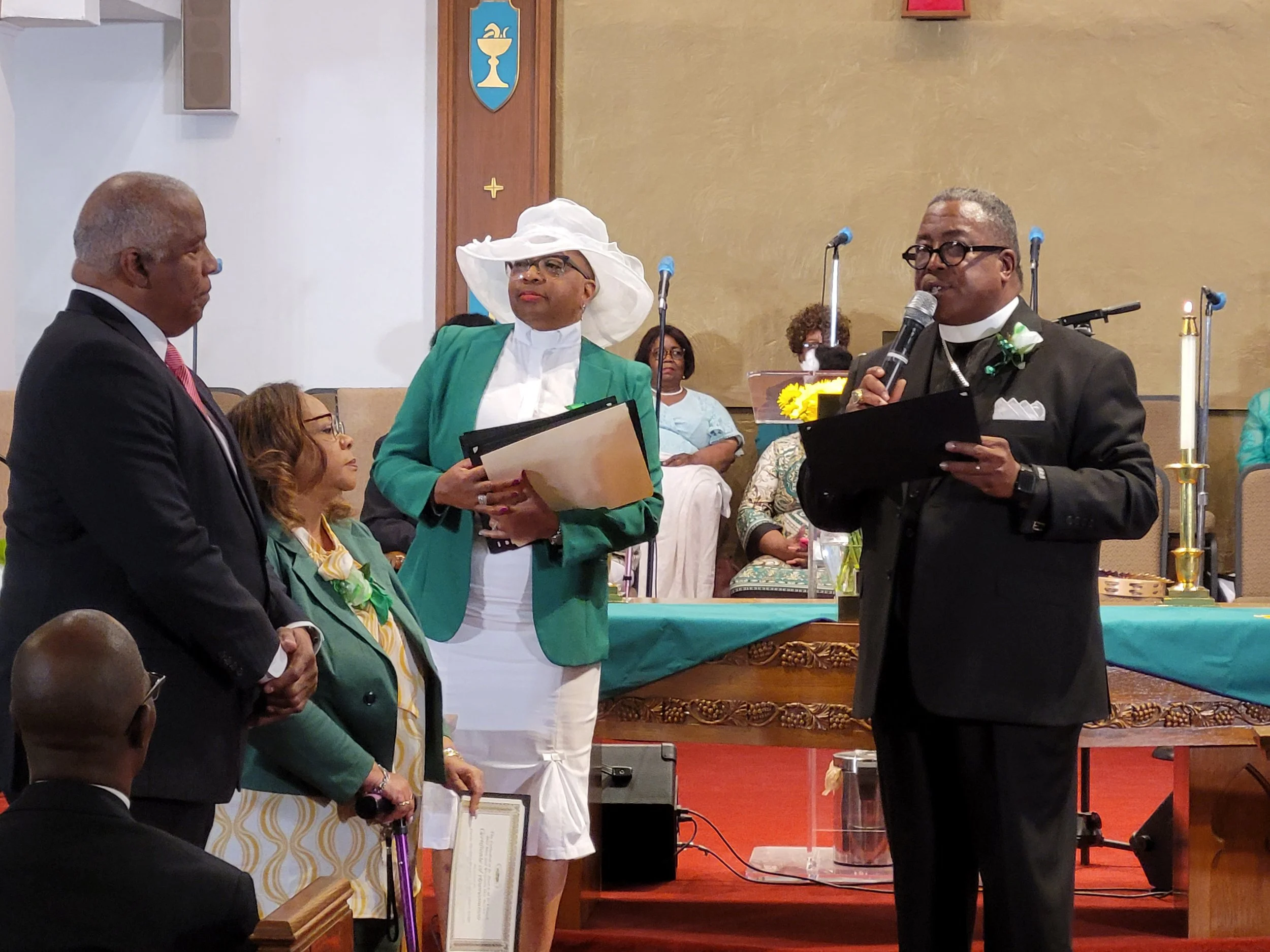 A church ceremony with a man in a black clerical suit speaking into a microphone, a woman in a white dress and large hat holding a folder, and a man in a suit with a pink tie listening. Several people are seated and standing in the background.