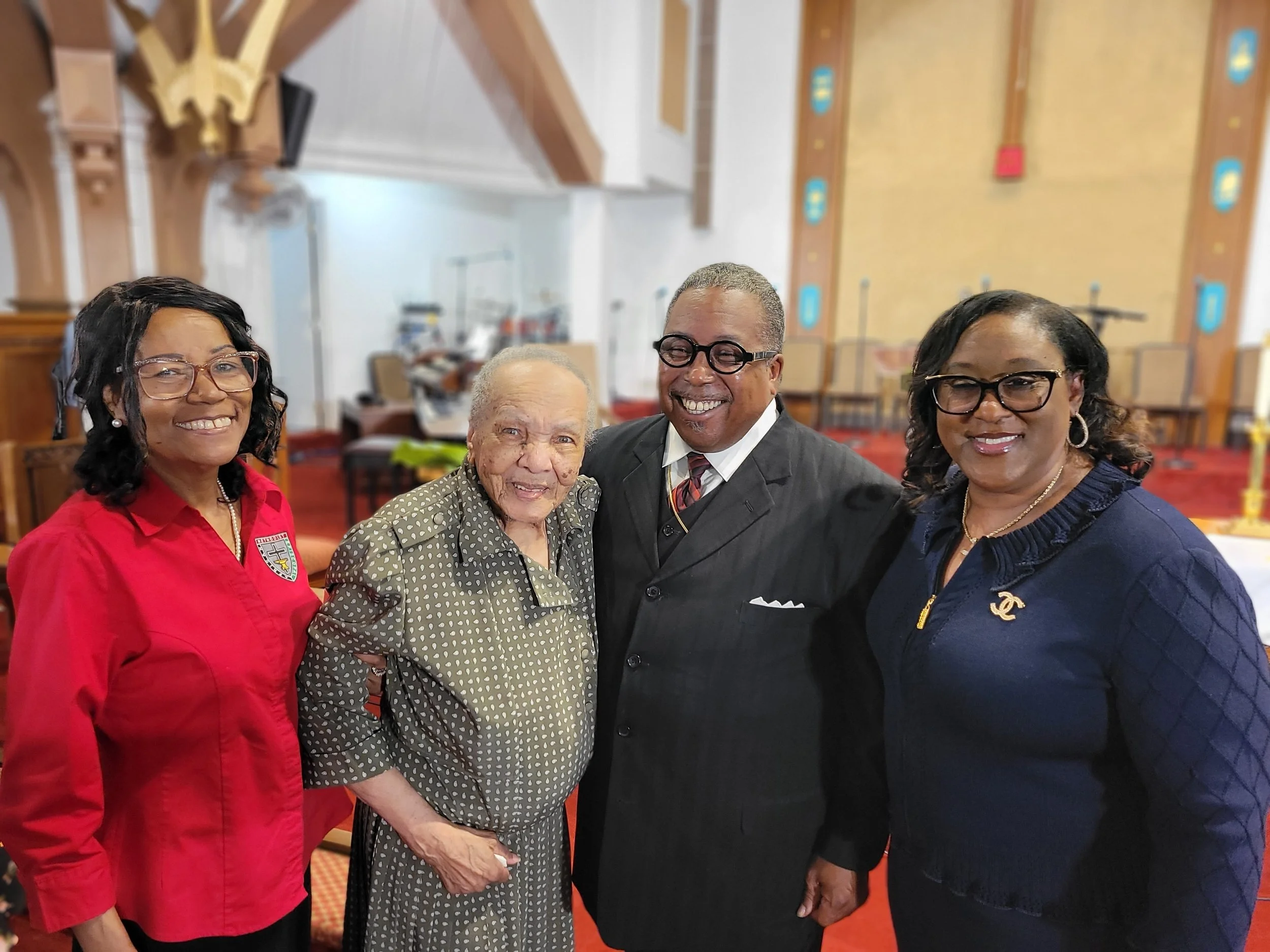 Four people smiling and standing together inside a church or similar building, with a woman who appears elderly in the center and three adults surrounding her, all dressed in semi-formal clothing.