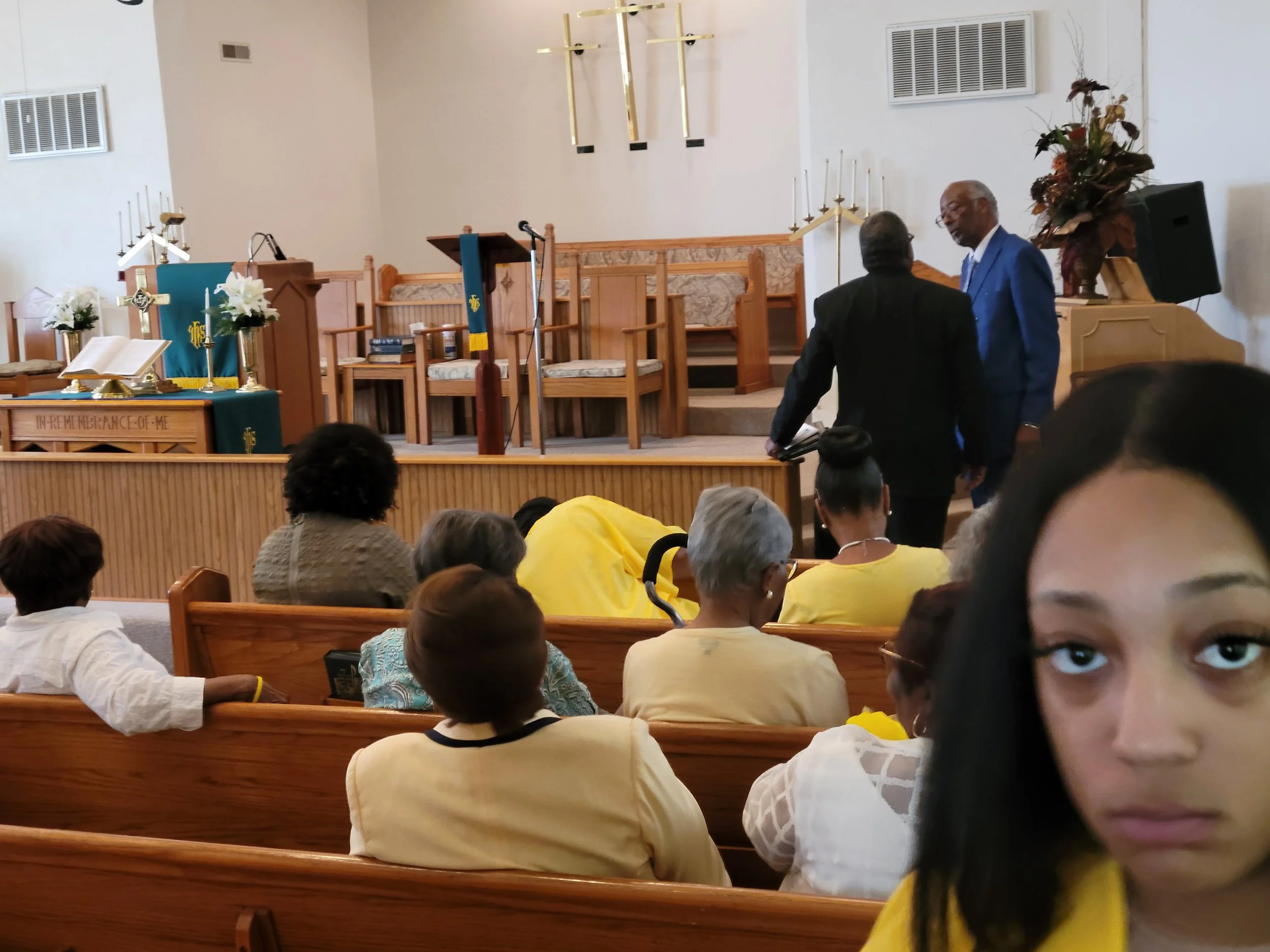 People gathered inside a church, some seated on wooden pews, with a platform at the front that has religious symbols, flowers, and an open Bible. Two men are standing and talking near the platform.