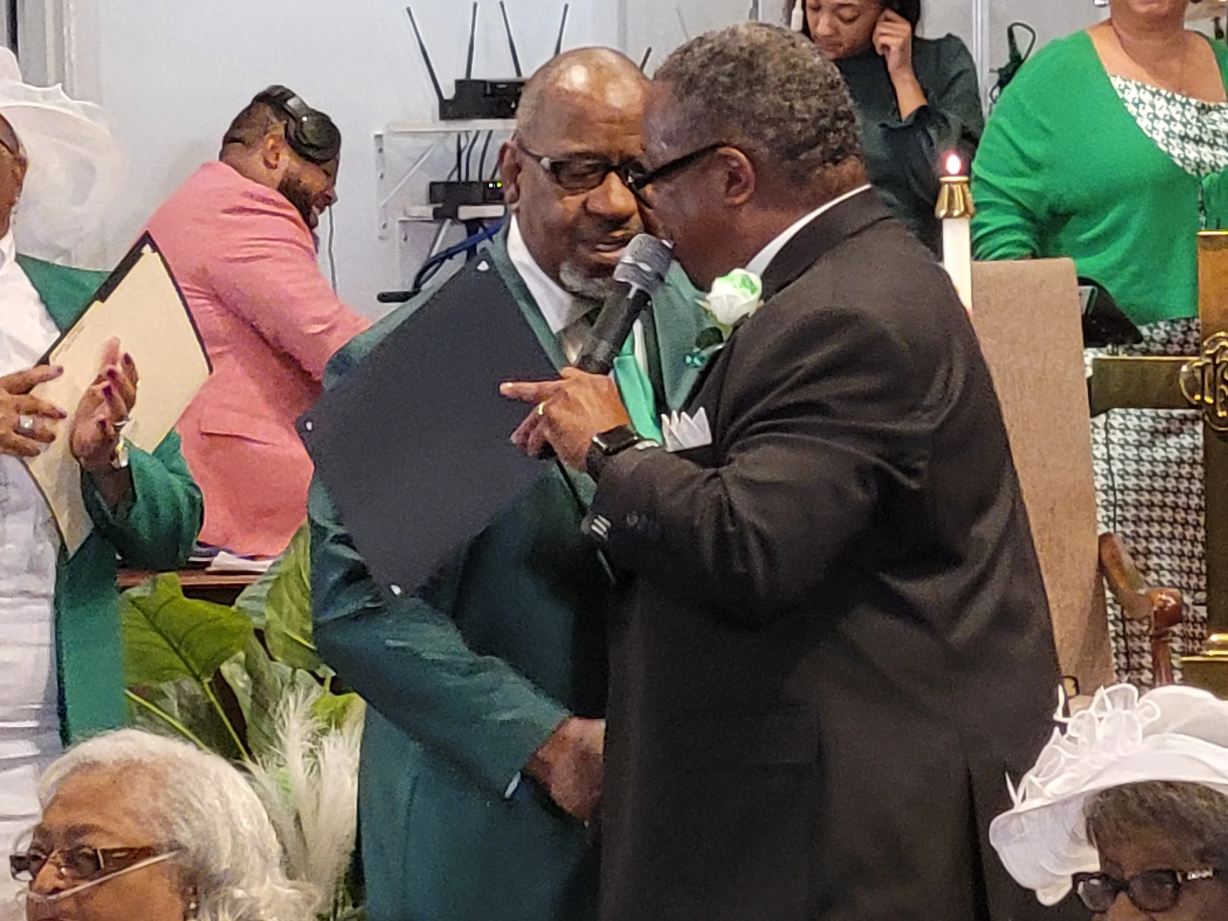 Two men are engaged in a close conversation, one holding a microphone and a black folder, both dressed in suits. The scene appears to be at an indoor formal event with several other people in the background, some standing and some seated, and various