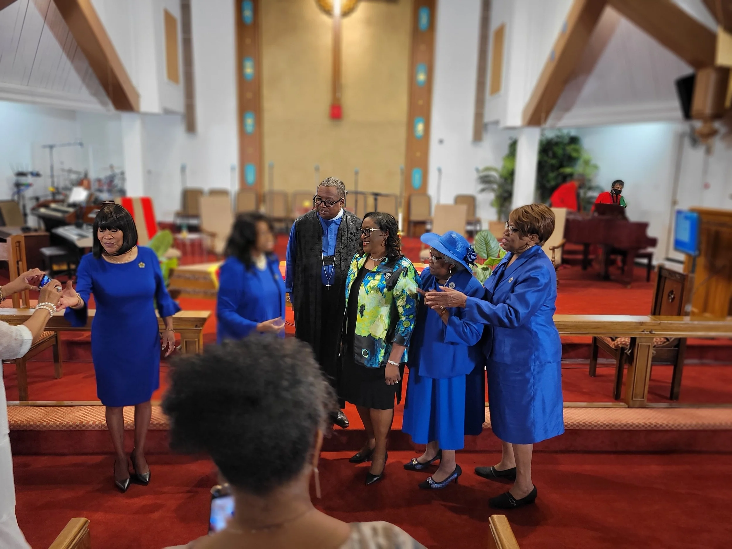 Group of people standing together in a church, dressed in formal attire, with a woman taking photos and a man playing the piano in the background.