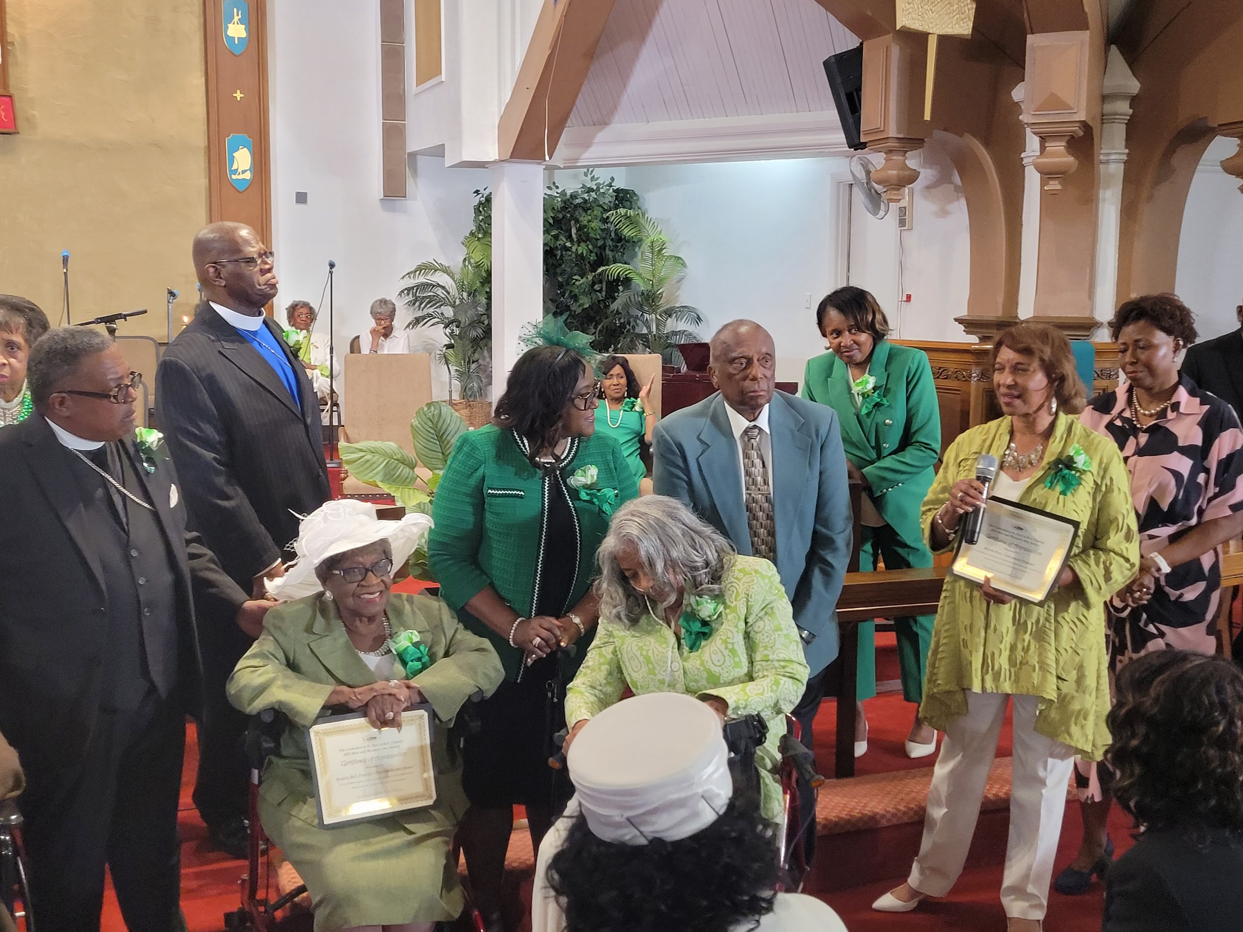 Group of people, including elderly women, gathered in a church for an award or certification ceremony, with a woman speaking into a microphone.