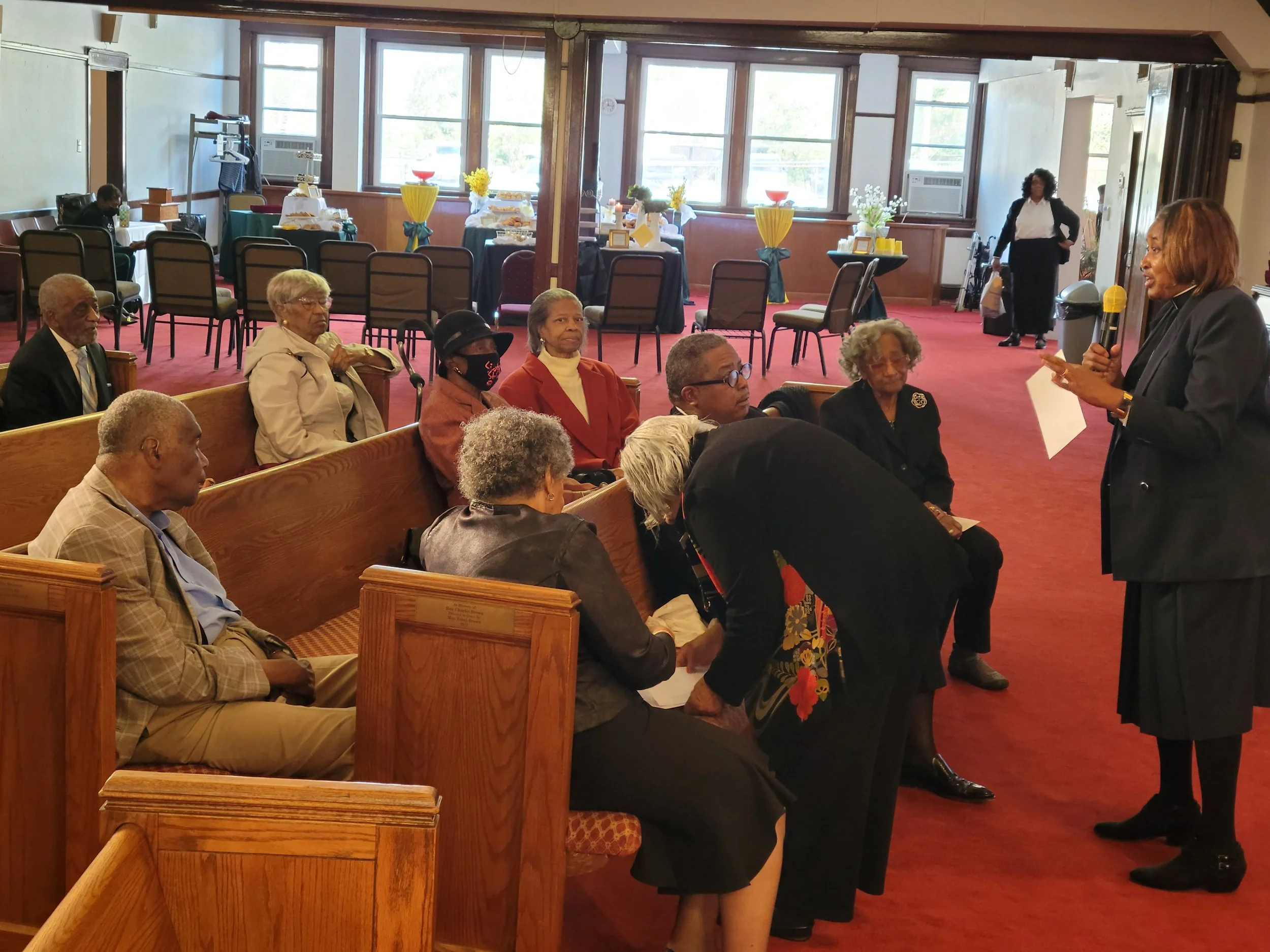A woman in a gray blazer and black dress holds a microphone and speaks to a seated group of seniors in a room with red carpet and decorated tables in the background.