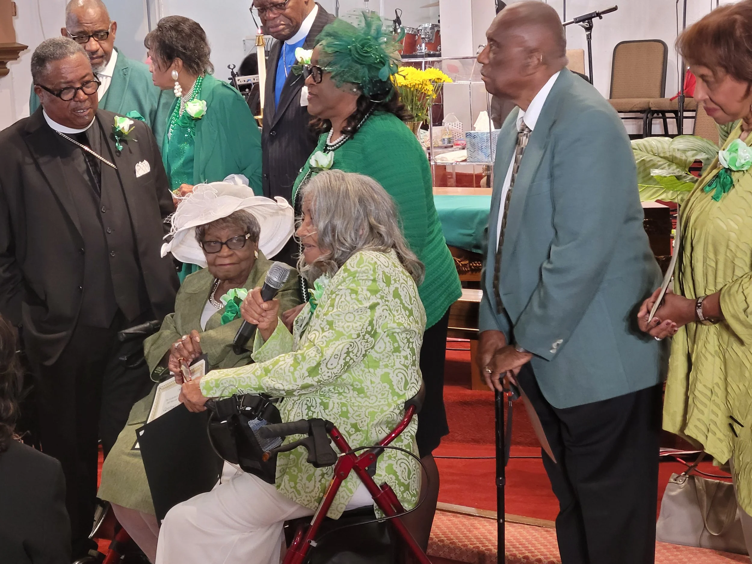 Group of elderly adults dressed in green, yellow, and formal attire, gathered around a seated woman with glasses and a large white hat, who is speaking into a microphone at a church or gathering event.