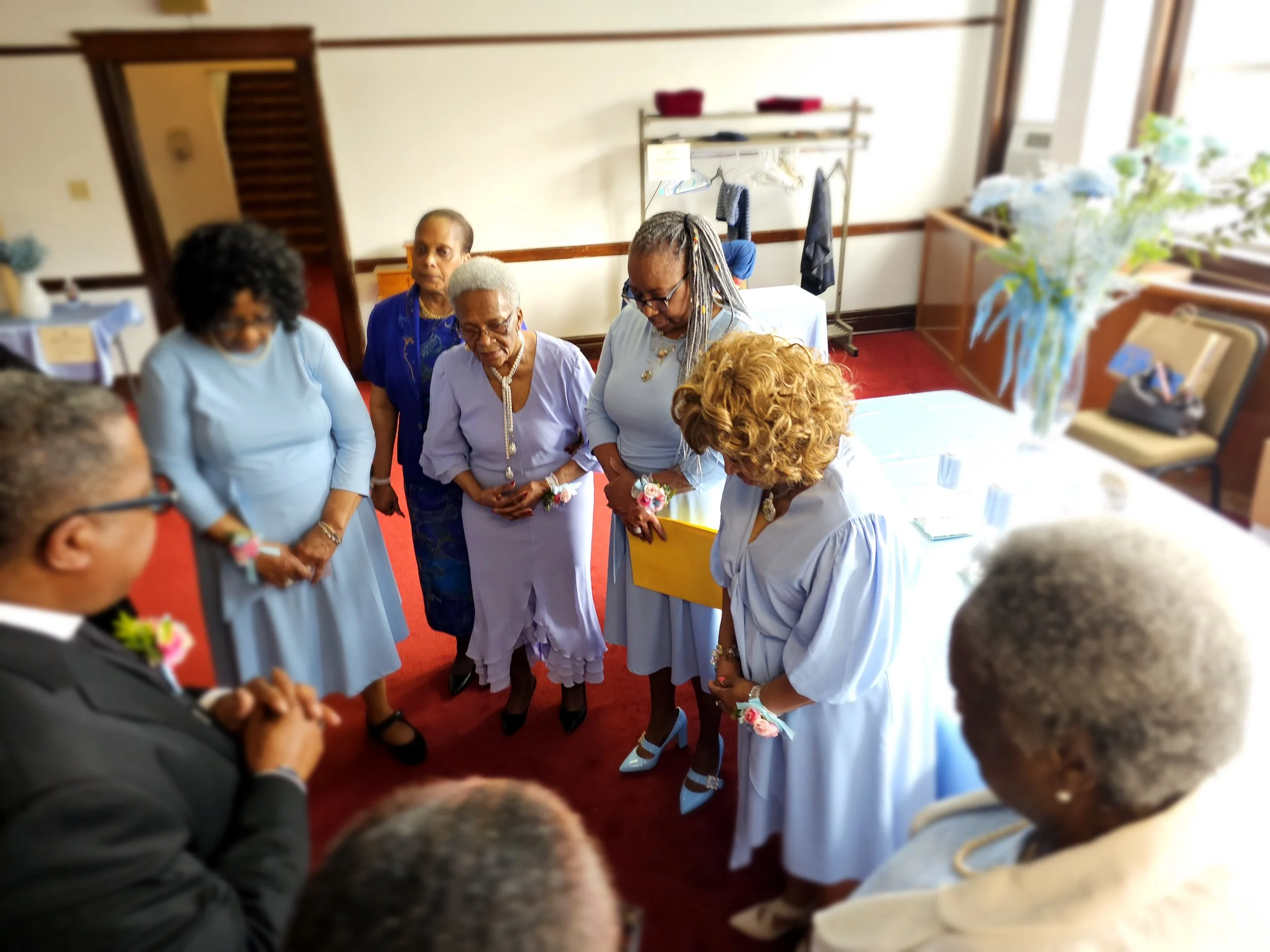 A group of elderly women and one man dressed in formal attire are gathered in a small room, apparently for a prayer or blessing. They are standing in a circle with their heads bowed and eyes closed, with a woman in a white dress holding a yellow fold