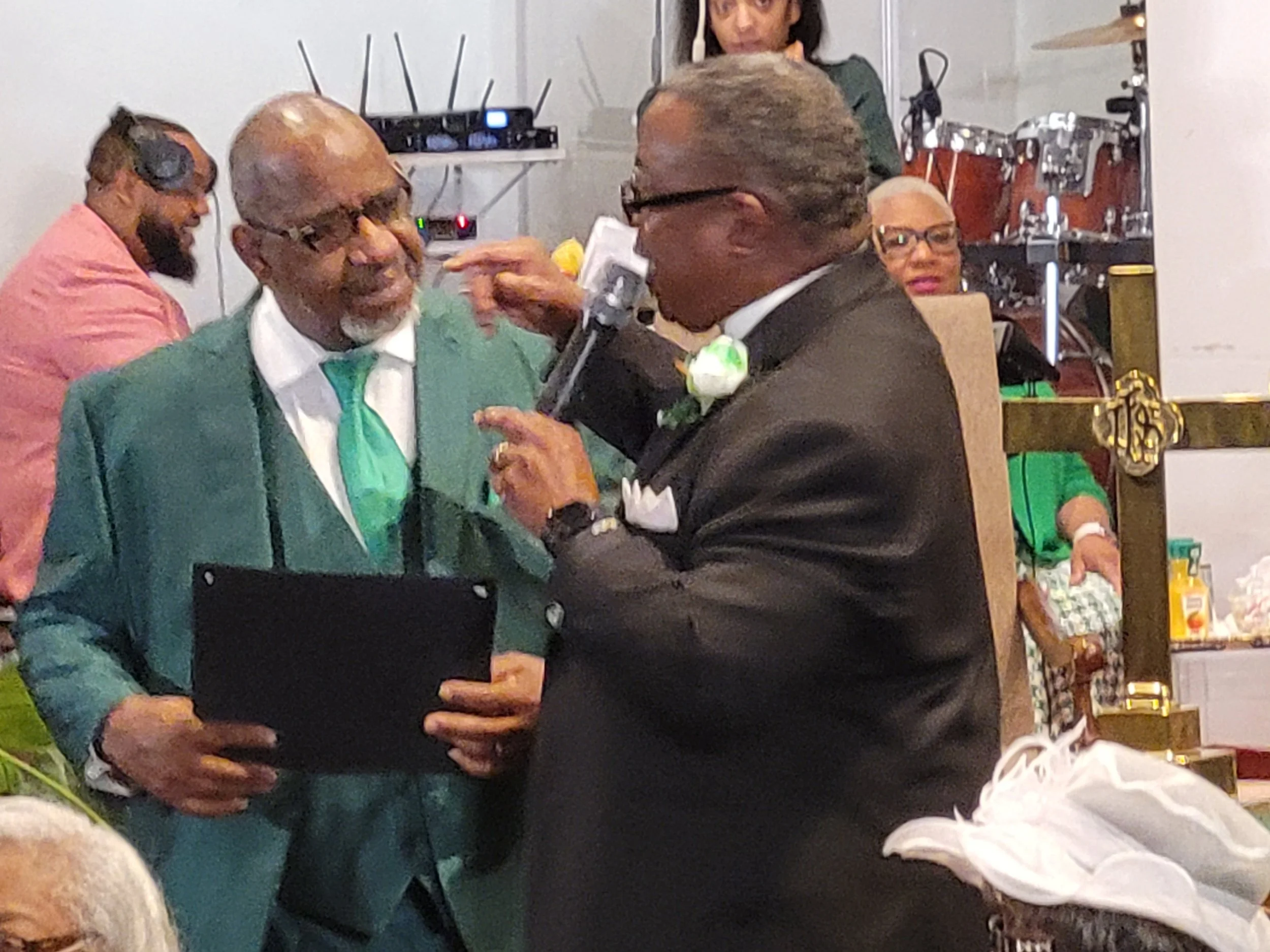 Two men celebrating at a wedding ceremony, one in a teal suit and the other in a black tuxedo, sharing a joyful moment with a microphone in hand.