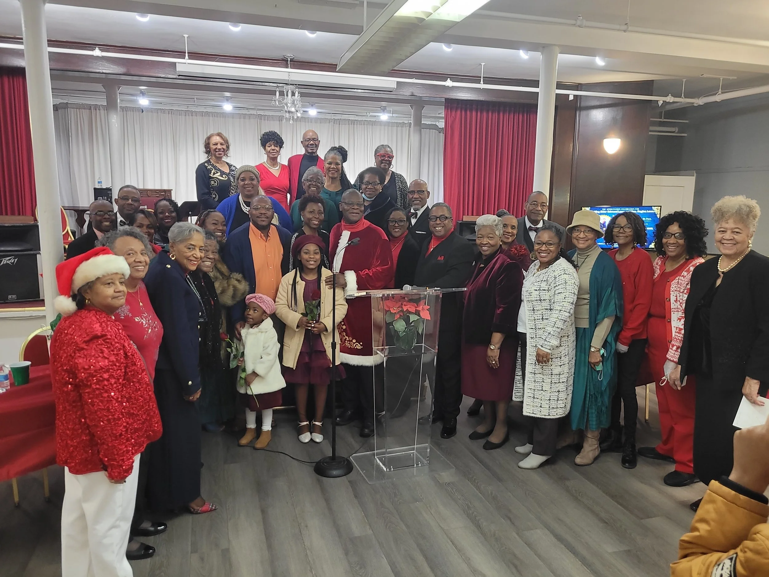 Group of people gathered indoors for a holiday celebration, with some wearing Christmas-themed clothing and accessories, including a woman in a Santa hat and a man dressed as Santa Claus, standing around a clear podium with a poinsettia plant.