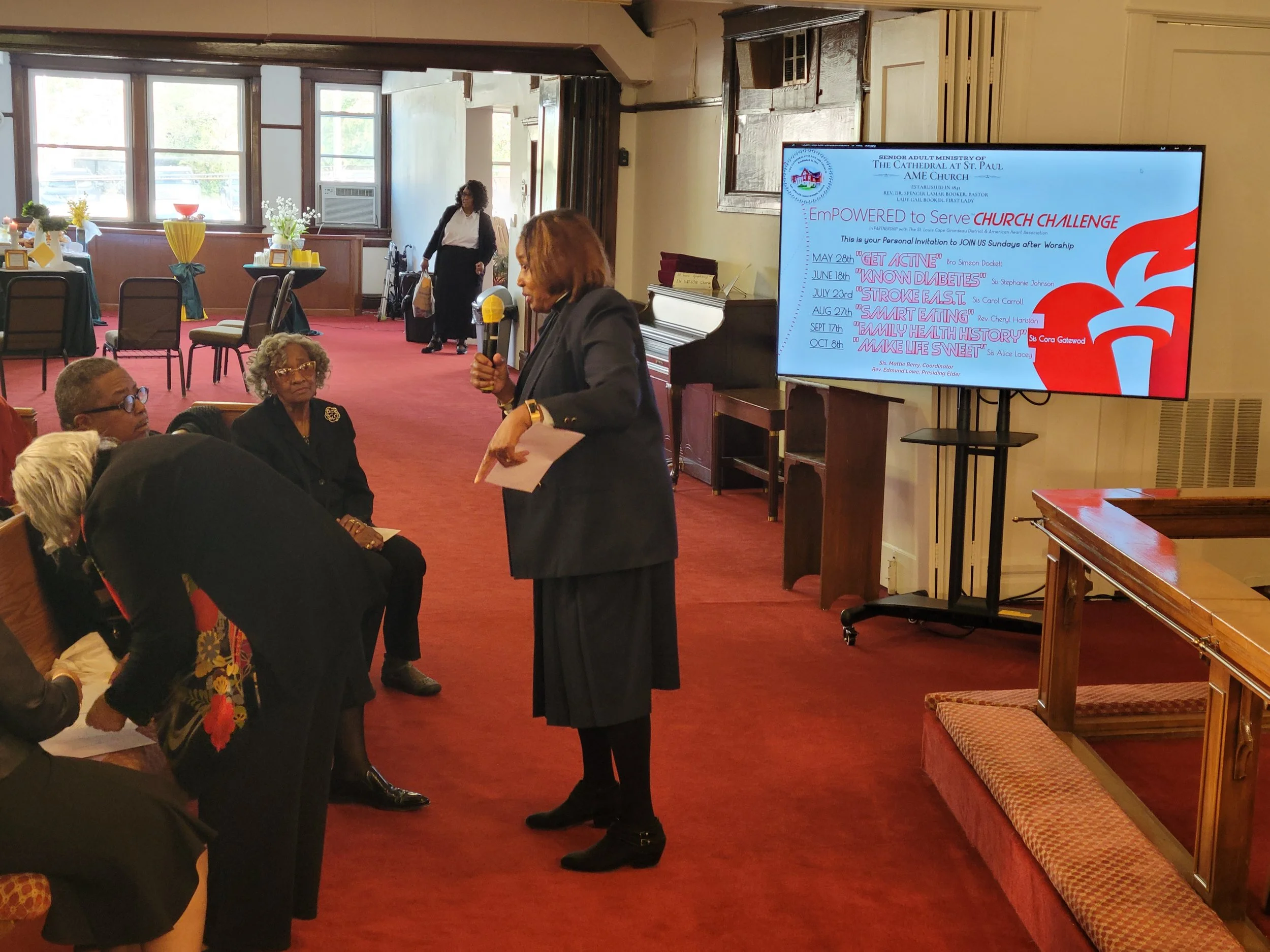 A woman standing with a microphone talking to a group of people in a room with red carpet and large windows. Behind her, a large screen displays a church event schedule.