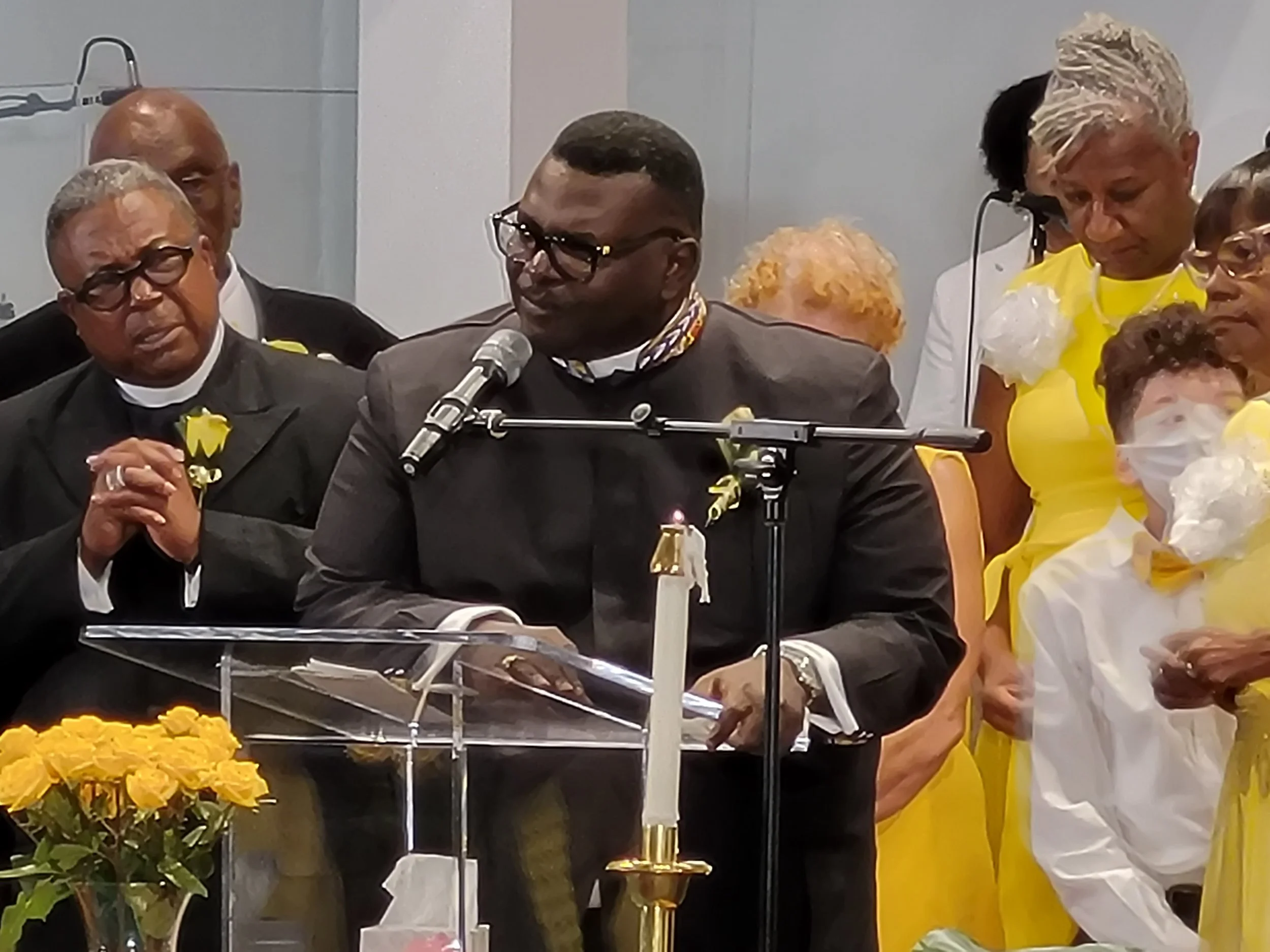 A group of people, including a man at a podium and others dressed in yellow, attending a ceremony with candles and flowers.